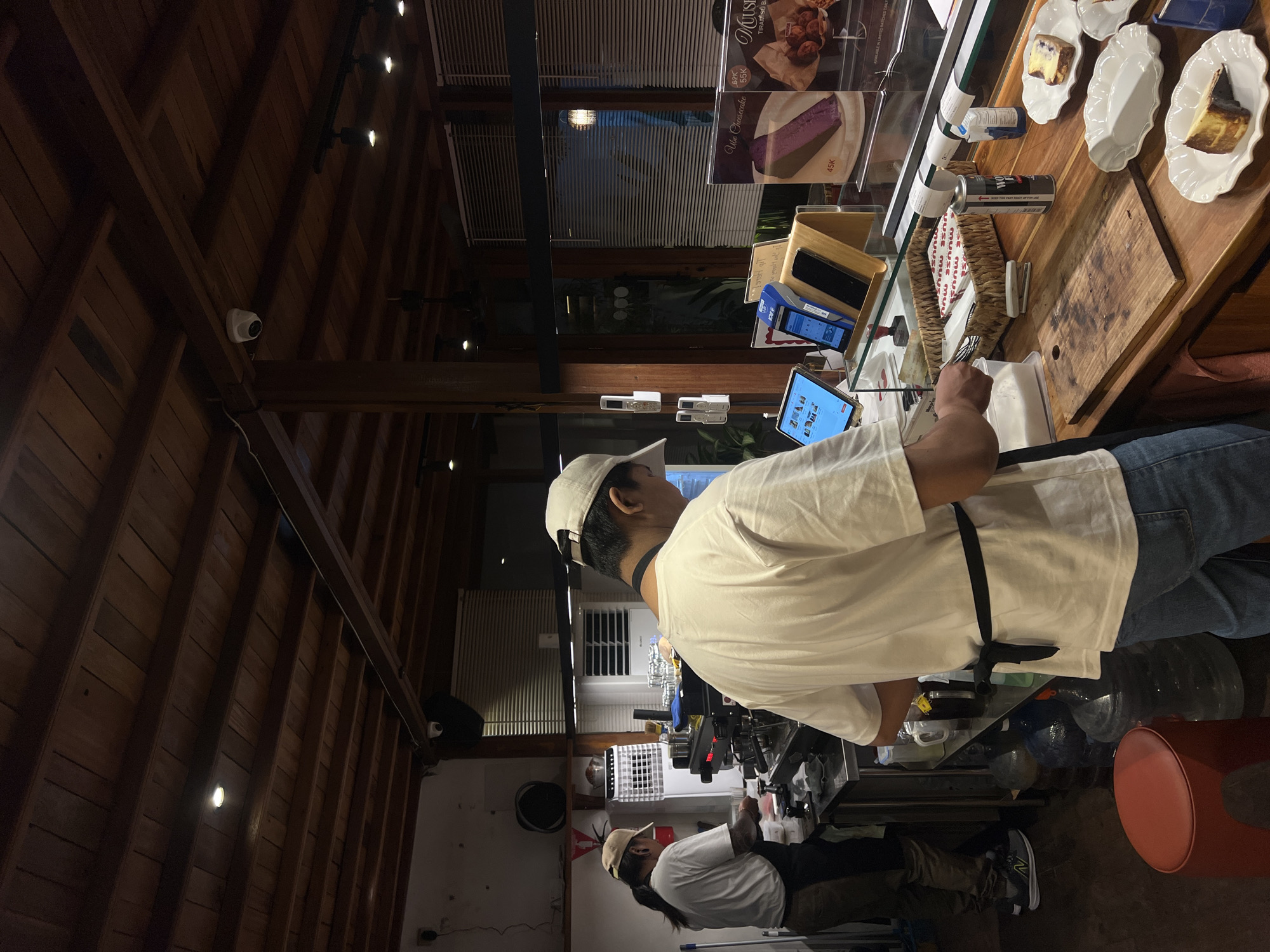 This image depicts the interior of a cafe in Mengwi, Indonesia. Two individuals, presumably cafe staff, are visible behind a counter. The person on the left, wearing a light-colored cap and apron, is preparing something at a coffee machine. The person on the right, facing away from the camera and wearing a white cap and t-shirt, appears to be engaged in a task at the counter, possibly serving or preparing food. The environment features a wooden ceiling with exposed beams and track lighting. Various objects are present, including a commercial coffee machine, tablets, payment terminals, pastries on display, and large water jugs. The overall lighting suggests it is evening or indoors with artificial lighting.