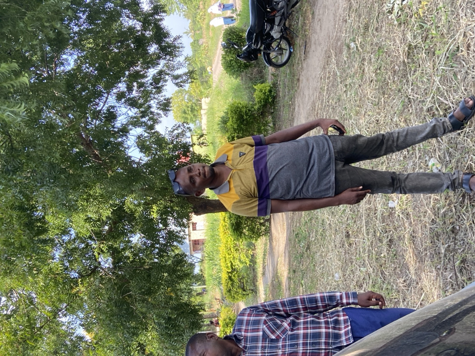 The image captures an outdoor scene in what appears to be a semi-urban or rural setting, likely in Kibaha, Tanzania, under bright daylight. The main subject is a man standing upright, facing slightly towards the camera, with a neutral expression. He wears a polo shirt with yellow shoulders and upper chest, a horizontal purple stripe across the mid-chest, and a grey lower torso. He has dark trousers and dark open-toed sandals. Sunglasses rest on top of his head, and a small dark object, possibly a phone, is held in his right hand.
To the left of the standing man, a large tree with dense green foliage dominates the background. A dirt path runs from the mid-ground towards the distant background, where a black motorcycle or scooter is parked. Beyond the path, there are green bushes and another, lighter-colored building with a reddish roof partially visible among the trees. In the far background, two distant figures, possibly in light-colored clothing, can be seen near a structure.
In the bottom right corner of the frame, another man is partially visible. He is wearing a plaid long-sleeved shirt with red, blue, and white squares, and dark blue trousers. Only his upper torso and head are visible, and he appears to be looking downwards, possibly leaning against or sitting in front of a dark-colored vehicle, a small part of which is visible in the extreme foreground. The ground is covered in dry grass and dirt. No text is visible in the image.