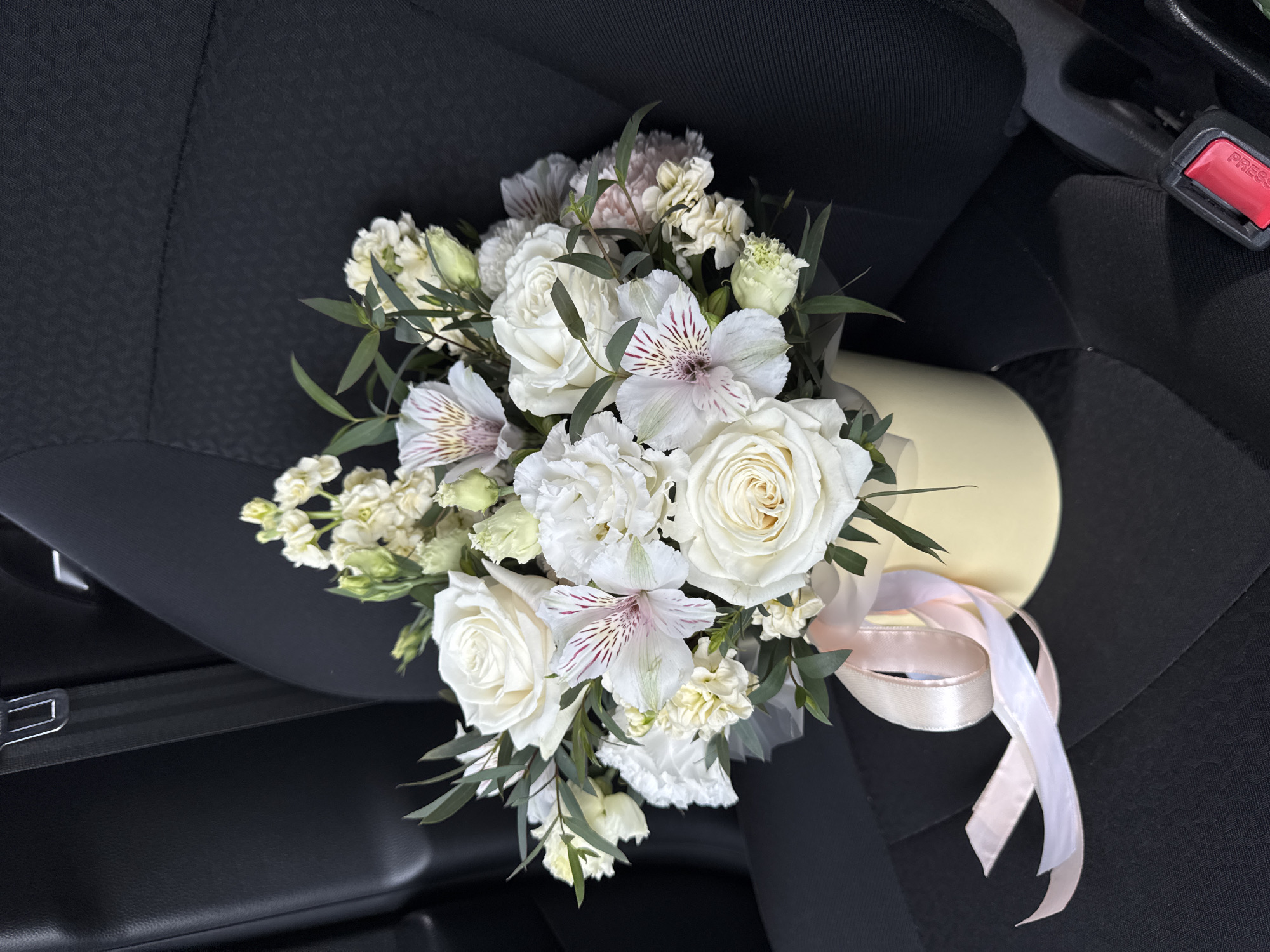 A floral arrangement rests on a black fabric passenger seat within an automobile interior. The bouquet is contained in a cylindrical, pale yellow hatbox. Pale pink and white satin ribbons are tied around the base of the hatbox, trailing onto the seat. The arrangement comprises a variety of white and cream-colored flowers, including several white roses, white alstroemeria with prominent pink and red internal striations, white eustoma (lisianthus), and clusters of white stock (Matthiola). Dark green, slender foliage is interspersed throughout the flowers. A black seatbelt is visible, with its silver buckle inserted into a receiver in the upper left of the frame and the red 'PRESS' release button of the buckle mechanism visible on the lower right. The car seat fabric exhibits a subtle textured pattern. No visual identifiers within the image provide information to confirm the specified geographic location of Izhevsk, Russia.
