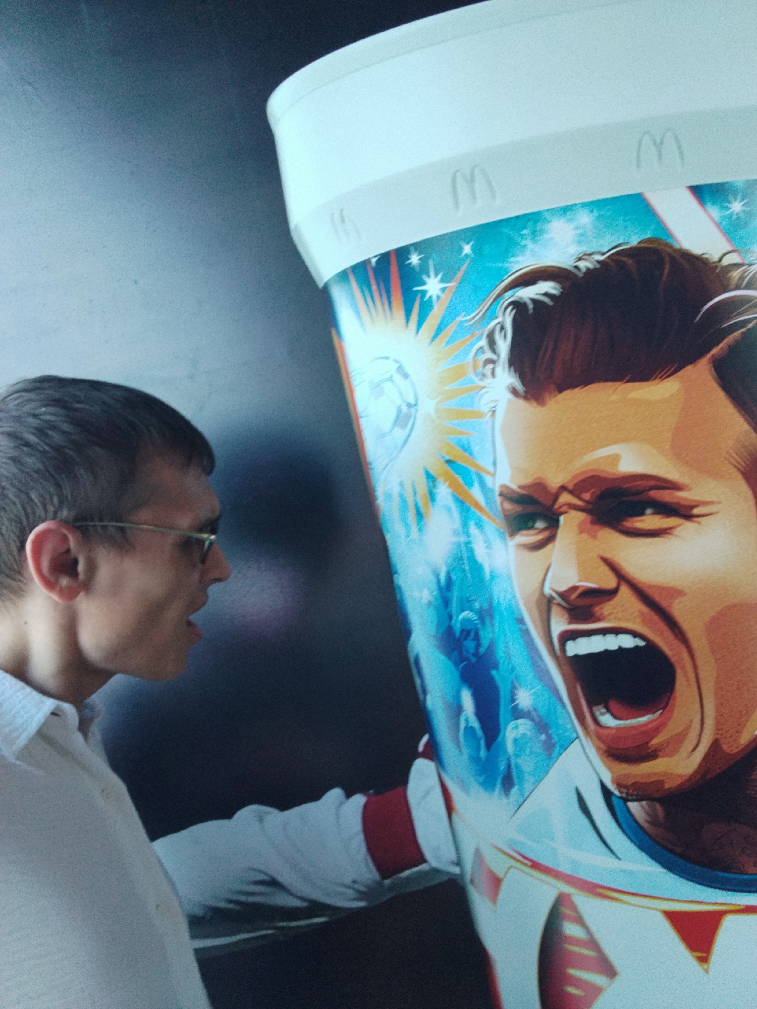 In Be'er Sheva, Israel, a man wearing glasses and a white shirt stands to the left of a large, cylindrical object adorned with a graphic of a man's face with his mouth wide open, reminiscent of a marketing display. The graphic also features a soccer ball and a starburst pattern, with subtle McDonald's logos visible on the top rim of the object. The man appears to be looking at the graphic, and his mouth is also slightly open, possibly in surprise or engaged in conversation. The background is a dark, textured wall. The lighting suggests an indoor environment, possibly a public space or a venue where such promotional material would be displayed.