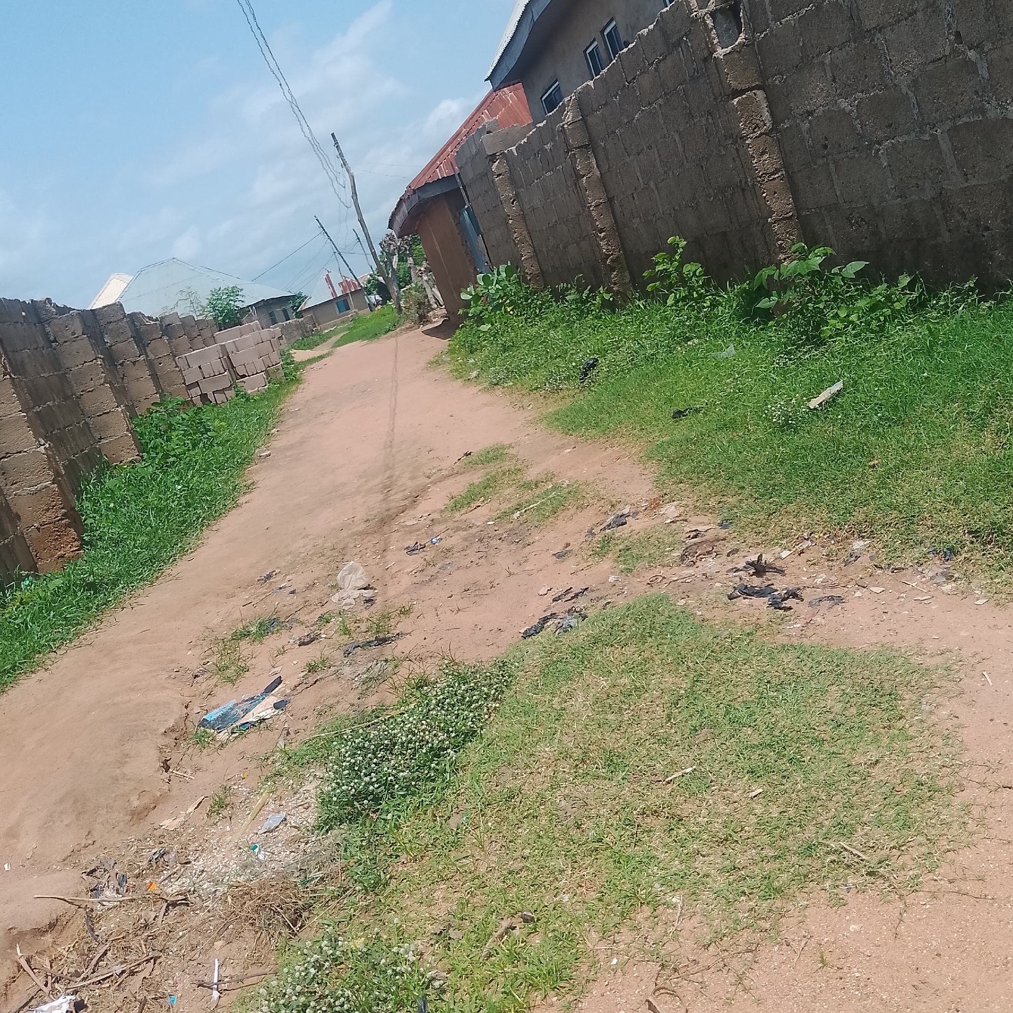 The image shows a dirt road in Jalingo, Nigeria, lined with cinder block walls and overgrown grass. The road surface is uneven and dusty, with scattered debris including plastic bags and other refuse. To the left, stacks of cinder blocks suggest ongoing construction or materials storage. Buildings with corrugated metal roofs are visible in the background, along with power lines. The sky is partly cloudy. The scene depicts a somewhat neglected urban or semi-urban environment.