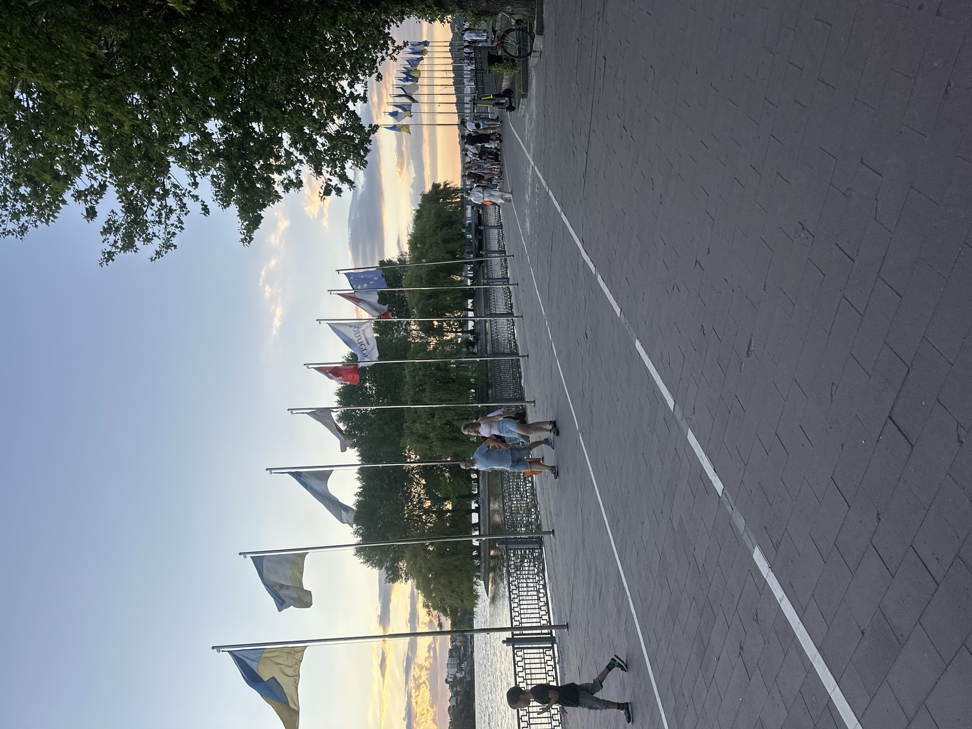 This is a horizontal, medium-shot, eye-level photograph taken at the Ternopil Lake embankment in Ternopil, Ukraine, during sunset. The embankment is paved with dark gray rectangular bricks and marked with two white dashed lines. On the left, a body of water, Ternopil Lake, is visible, with trees and buildings on the opposite shore. A row of flags on metal poles stands along the embankment, with the Ukrainian flag prominently displayed on the left. Several people are walking or standing along the embankment. On the left, a young boy in a black t-shirt and gray shorts is walking towards the right. A couple, a man in a blue t-shirt and shorts and a woman in a pink tank top and white shorts, are walking together towards the right. Further down the embankment, a larger group of people is congregated, some walking and others standing, with a few e-scooters and a bicycle visible. The sky is a soft gradient of blue and orange, with some clouds indicating the setting sun. The overall scene depicts a peaceful evening stroll along the waterfront.