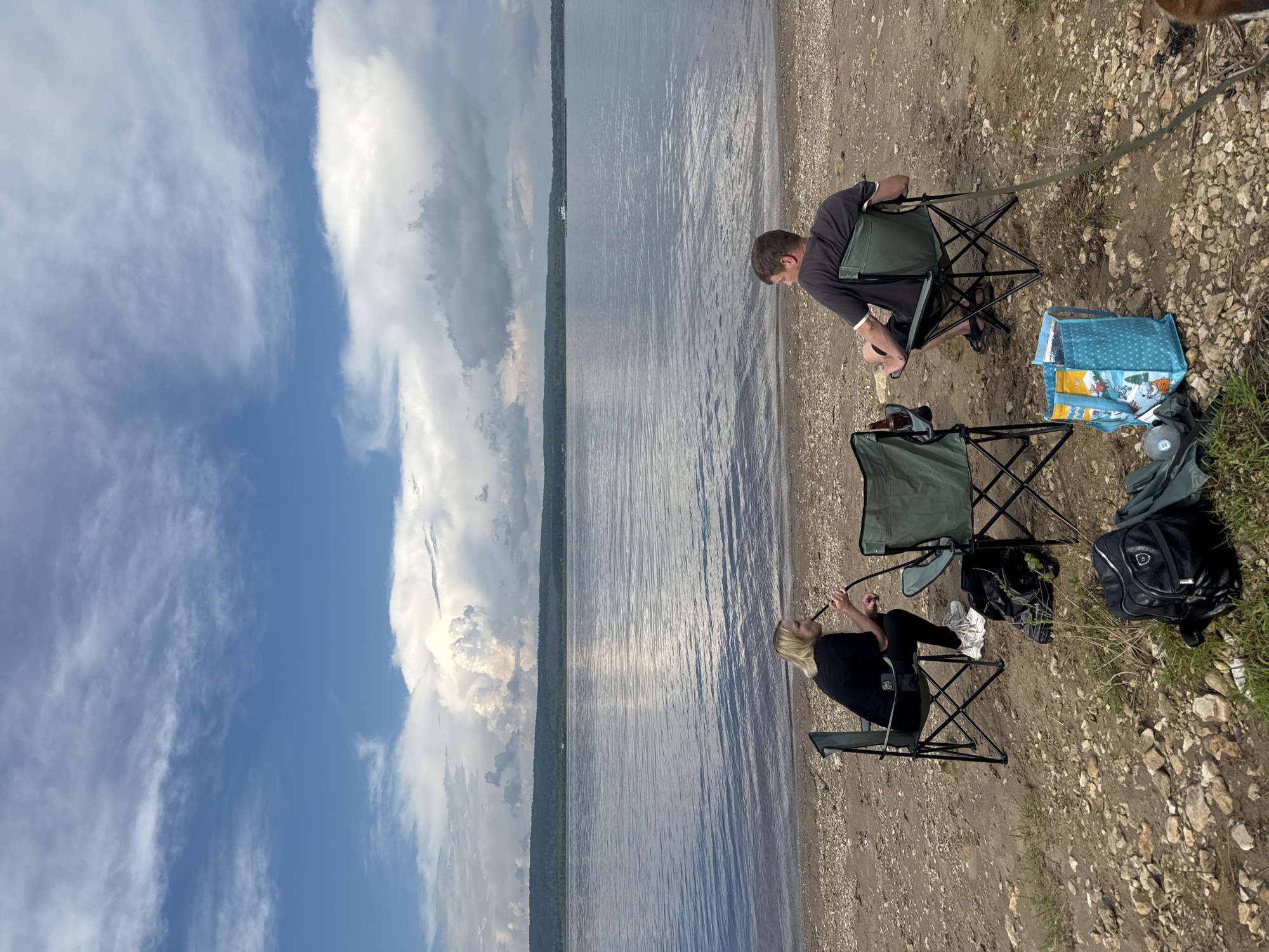 Two people are sitting on camping chairs on a rocky shore by a wide body of water. A woman with blonde hair is on the left, wearing a black shirt and pants, and appears to be smoking a hookah. A man with brown hair is on the right, wearing a grey t-shirt and dark shorts. He is looking away from the camera, towards the water. There are two green camping chairs, a black backpack, a blue patterned bag, and a dark bag on the ground. The water stretches to a distant tree-lined shore under a sky with dramatic clouds.