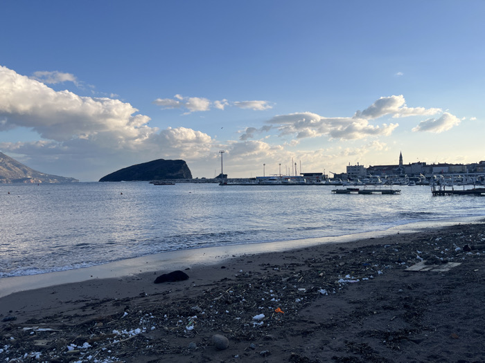 The image depicts a coastal scene in Budva, Montenegro, looking across the bay towards Sveti Nikola Island and the Old Town.
The foreground consists of a dark pebble and sand beach heavily strewn with debris. This debris includes numerous pieces of white and dark plastic, wood fragments, and light-colored unidentifiable refuse. A narrow strip of wet sand marks the edge where small, rippled waves are breaking. A dark, irregularly shaped object rests on the wet sand near the water's edge. No individuals or animals are present on the beach.
The middle ground displays the calm, grey-blue water of the Adriatic Sea. In the center-left, the dark, vegetated mass of Sveti Nikola Island is prominent on the horizon. To the right, a marina is visible, populated by multiple white-hulled recreational boats moored at various floating docks and piers. A tall, slender light pole or mast stands at the marina's entrance.
The background on the far right reveals the historic skyline of Budva Old Town, characterized by densely packed stone buildings with red-tiled roofs and a distinct, pointed bell tower, identifiable as St. Ivan Church. Distant, hazy mountains rise along the far left horizon. The sky is predominantly clear blue with scattered, fluffy white cumulus clouds, illuminated by soft light from the right, suggesting late afternoon.
