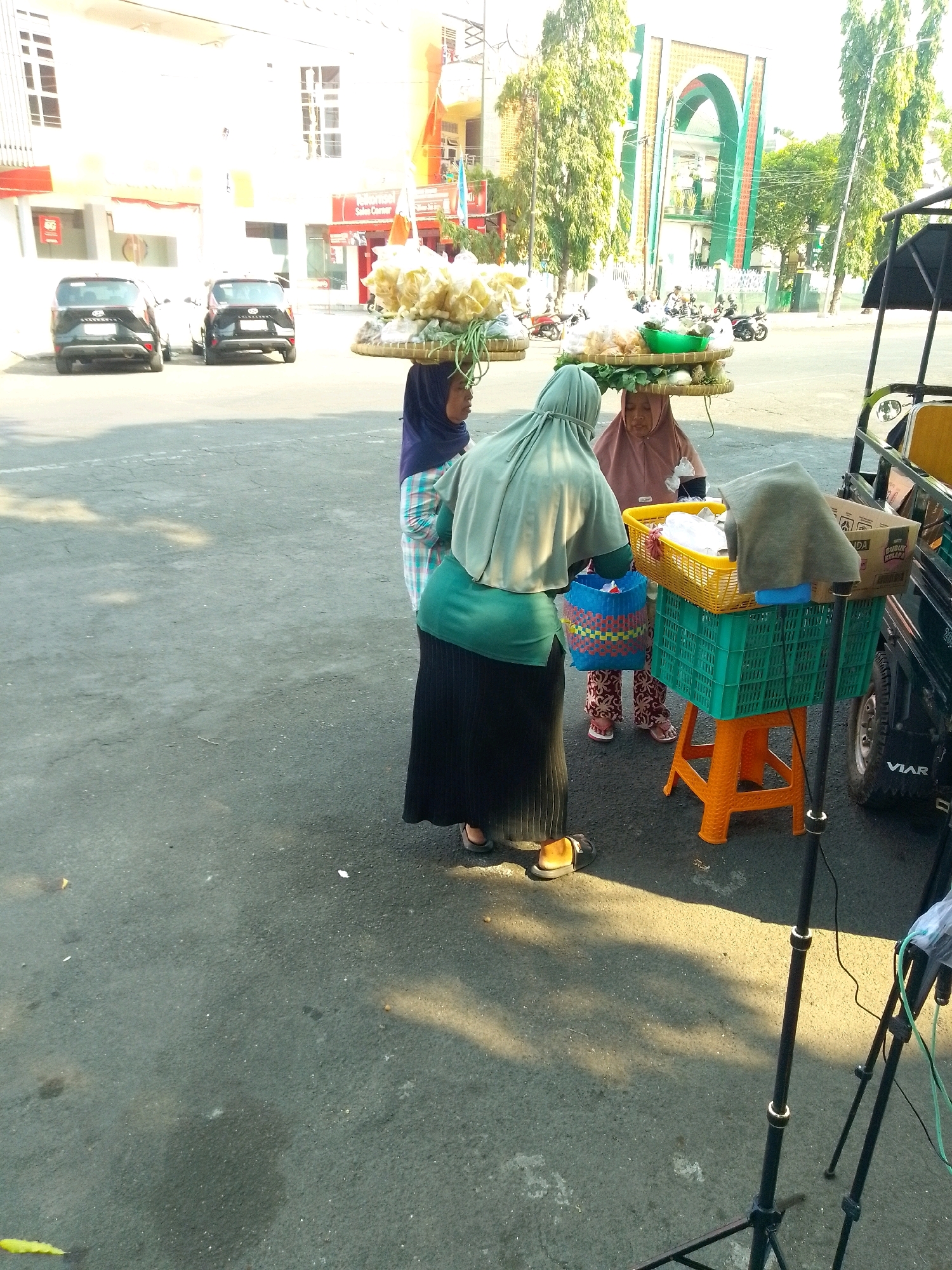 The image depicts a street scene in Jember, Indonesia, possibly near a mosque given the architectural style of the building in the background. In the foreground, three women are engaged in a transaction, likely selling food. The woman on the left wears a blue and white checkered shirt and a dark blue hijab, carrying a woven basket on her head. The woman in the center, wearing a light green hijab and a black skirt, is facing the woman on the right and appears to be handling a blue woven bag. The woman on the right, in a pink hijab, is also interacting with the woman in the center.
In front of the women, a yellow basket is placed on top of a green plastic crate, which is supported by an orange plastic stool. The yellow basket contains various items, possibly food products. A grey cloth is draped over some of the contents of the yellow basket. Next to the baskets, a black vehicle with "VIAR" branding is visible, suggesting it might be a three-wheeled cargo vehicle used for transport. The vehicle is partially covered by a black canopy. A cardboard box with Indonesian text is also visible on the vehicle.
The ground is paved with asphalt, and sunlight casts distinct shadows, indicating it is daytime. In the background, two black cars are parked. Beyond the cars, a white building with multiple windows and signs is present. Further in the background, a building with an arched entrance, reminiscent of mosque architecture, is visible, surrounded by tall green trees. Several motorcycles are parked or in motion in the background, suggesting a busy urban environment. Two camera tripods with attached equipment are positioned in the lower right corner of the image, hinting at a possible filming or photography activity.