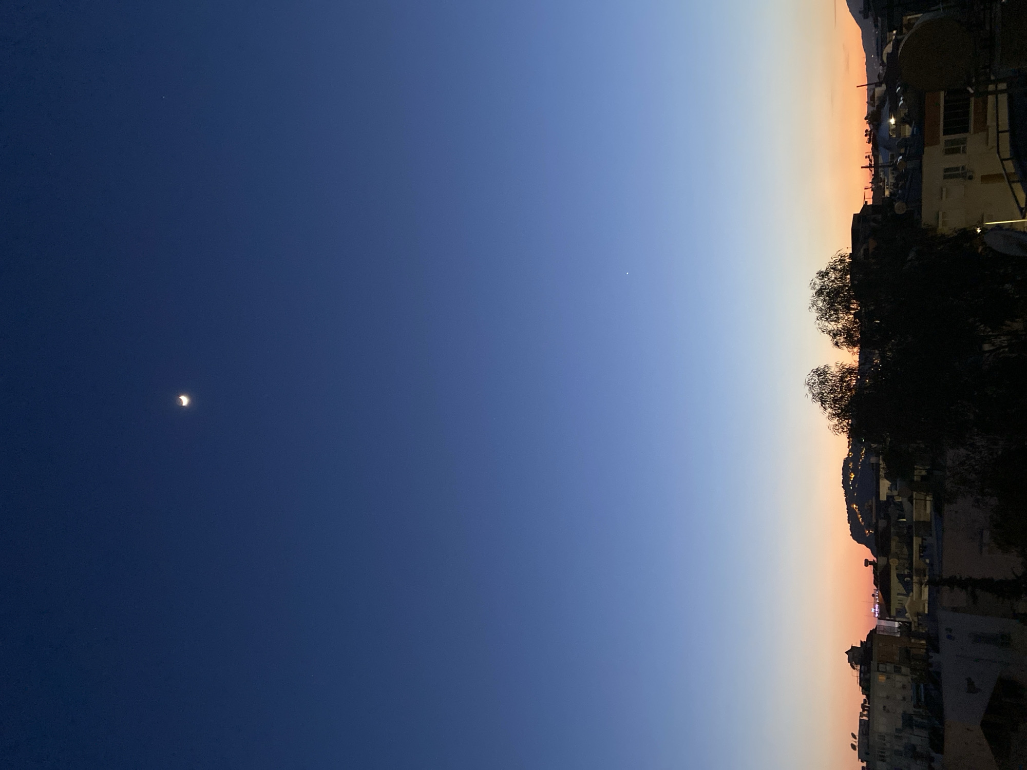 The image presents a tranquil twilight scene over Alanya, Turkey. The sky, occupying the majority of the frame, displays a striking horizontal gradient. It transitions from a deep, dark indigo blue on the far left, through a lighter, vibrant blue in the mid-section, to warm orange and pink hues along the right horizon.
In the deep blue portion of the sky on the left, a distinct, luminous crescent moon is visible. Further to the right, within the lighter blue expanse, a faint, bright white dot, likely a planet or a prominent star, can be discerned.
Along the bottom of the image, the dark silhouette of a city or town stretches across the horizon. Various buildings, appearing as dark shapes against the illuminated sky, suggest a mix of residential and commercial structures. Some faint lights emanating from within the city indicate that it is either dusk or dawn. Beyond the immediate urban structures, the dark outlines of hills or mountains rise in the background. Several dark, silhouetted trees are also visible, interspersed within the cityscape and in the foreground.
The time of day is clearly twilight, characterized by the dramatic color transition in the sky, from the deep hues of impending night to the softer glow of the setting or rising sun. The scene evokes a calm and serene atmosphere. There are no identifiable people, specific activities, or visible text in the image.