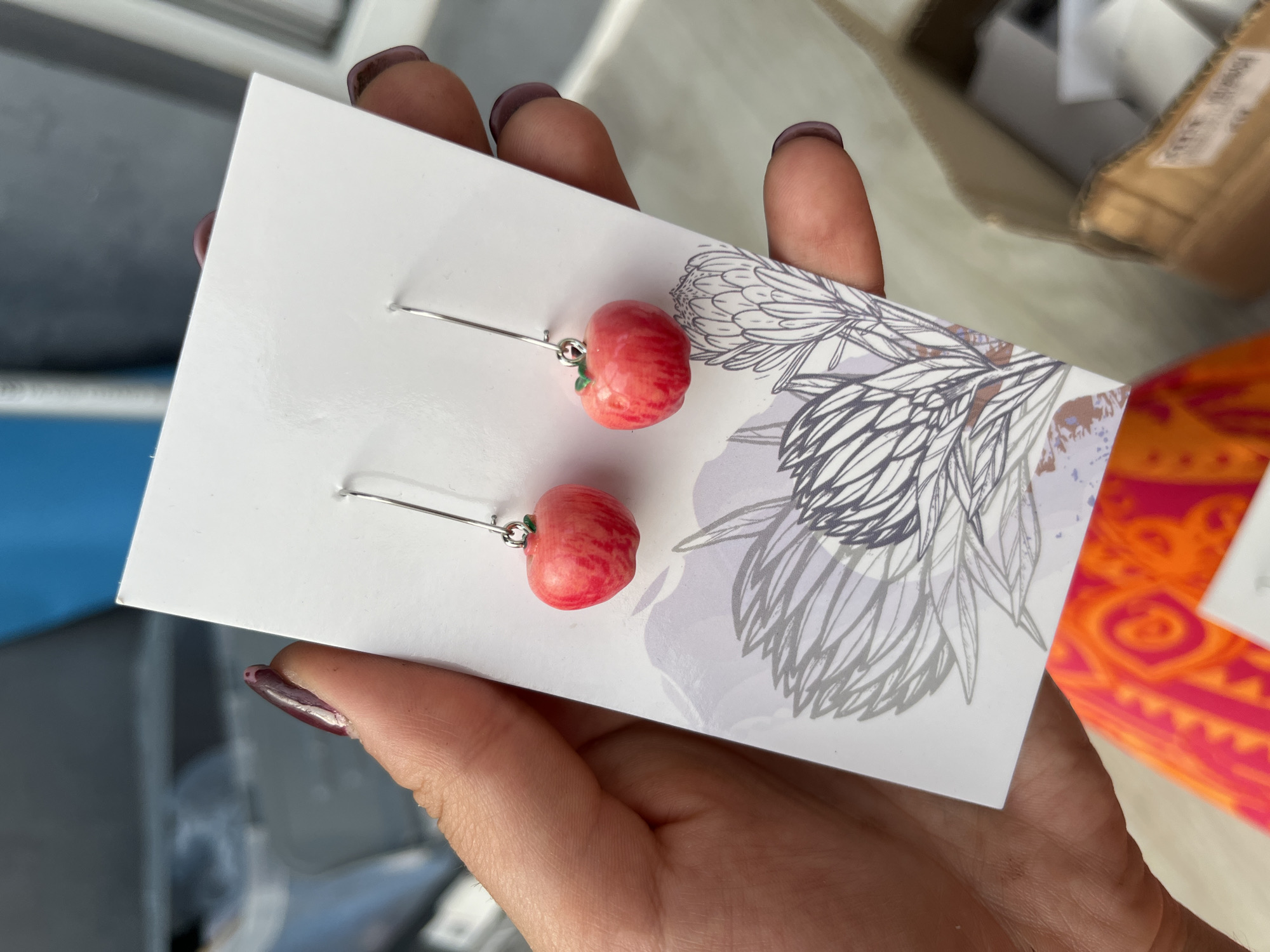 In Сеніцкі сельскі Савет, Belarus, a hand with dark nail polish holds a white card featuring a line drawing of a protea flower. Two dangle earrings, each shaped like a small, red apple with a green stem, are attached to the card. The background is out of focus, showing a blurred interior setting with shelves and boxes.