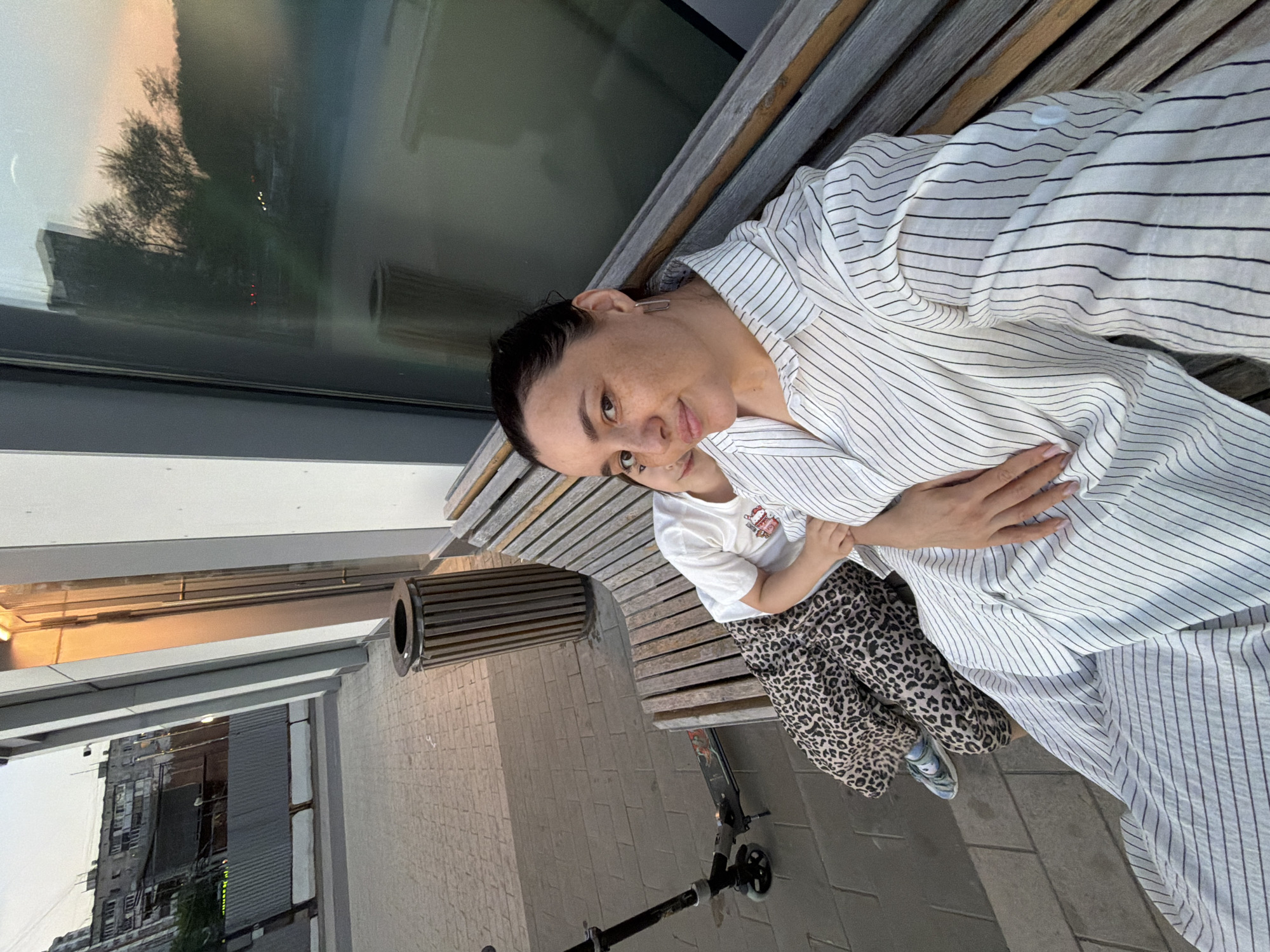 In Moscow, Russia, two individuals, an adult woman and a child, are seated on a wooden bench outside. The woman, positioned in the foreground, is taking a selfie, looking directly at the camera with a slight smile. She is wearing a white shirt with thin black vertical stripes and large hoop earrings. Her left hand rests on her lap. Beside her, a child is partially visible, leaning against her. The child is wearing a white t-shirt and patterned trousers. To the left of the bench, a scooter is parked on a paved walkway. In the background, a trash bin made of wooden slats is visible, along with the exterior of a building featuring large glass windows reflecting the surrounding environment and the sky. The architecture suggests an urban setting, likely a public space or commercial area.