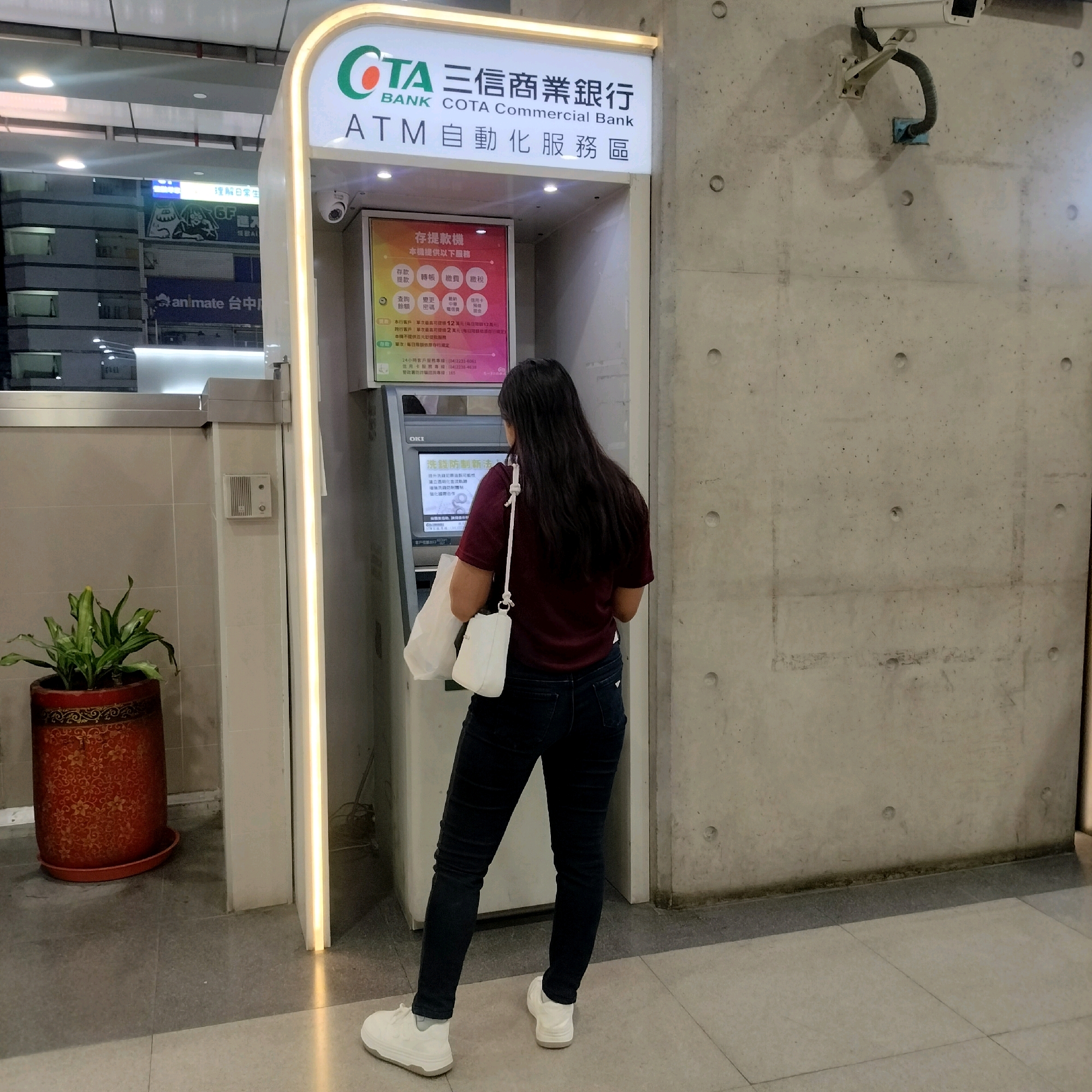 A woman with long, dark hair is standing in front of an ATM machine in Taichung, Taiwan. She is wearing a maroon t-shirt and dark jeans. A white purse is slung over her shoulder, and she is holding a white shopping bag in her left hand. The ATM is part of COTA Commercial Bank. To the left of the ATM is a potted plant. The background shows a concrete wall and a building with signage. The image appears to be taken indoors or in a covered area, possibly a financial institution or a public transportation hub.