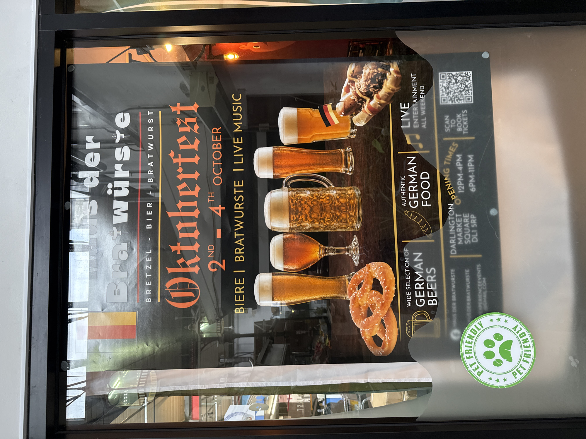 This image shows a window display for an Oktoberfest event hosted by "Haus der Bratwurst" in Darlington, United Kingdom. The poster features the German flag, text promoting "Bier | Bratwurst", and images of beers and pretzels. It advertises "Oktoberfest 2nd - 4th October" with "Authentic German Food", "Live Entertainment", and "Wide Selection of German Beers". A "Pet Friendly" sticker is affixed to the window. The lower section of the poster includes event details such as "Darlington Market Square DL1 5RP", "Opening Times 12PM-4PM" and "6PM-11PM", and a QR code for "Scan to Book Tickets". The reflection in the window shows the interior of a building with lights and shelves.