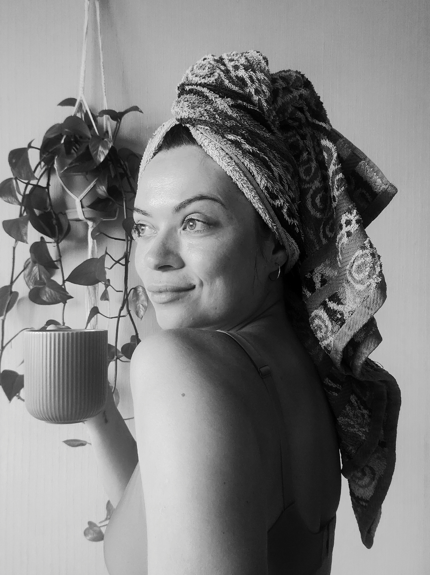 A black and white medium shot depicts a fair-skinned woman with a towel wrapped around her head. She is turned slightly to the left with her head turned back towards the viewer, and she is smiling gently while looking towards the left. She is holding a ribbed, cylindrical object, possibly a mug or a pot, in her left hand. Her right arm is visible, and she appears to be wearing a tank top or similar garment. A hanging plant is visible behind her, to the left. The background is a plain wall. No information confirms the location in the city of Chaikovsky, Russia.