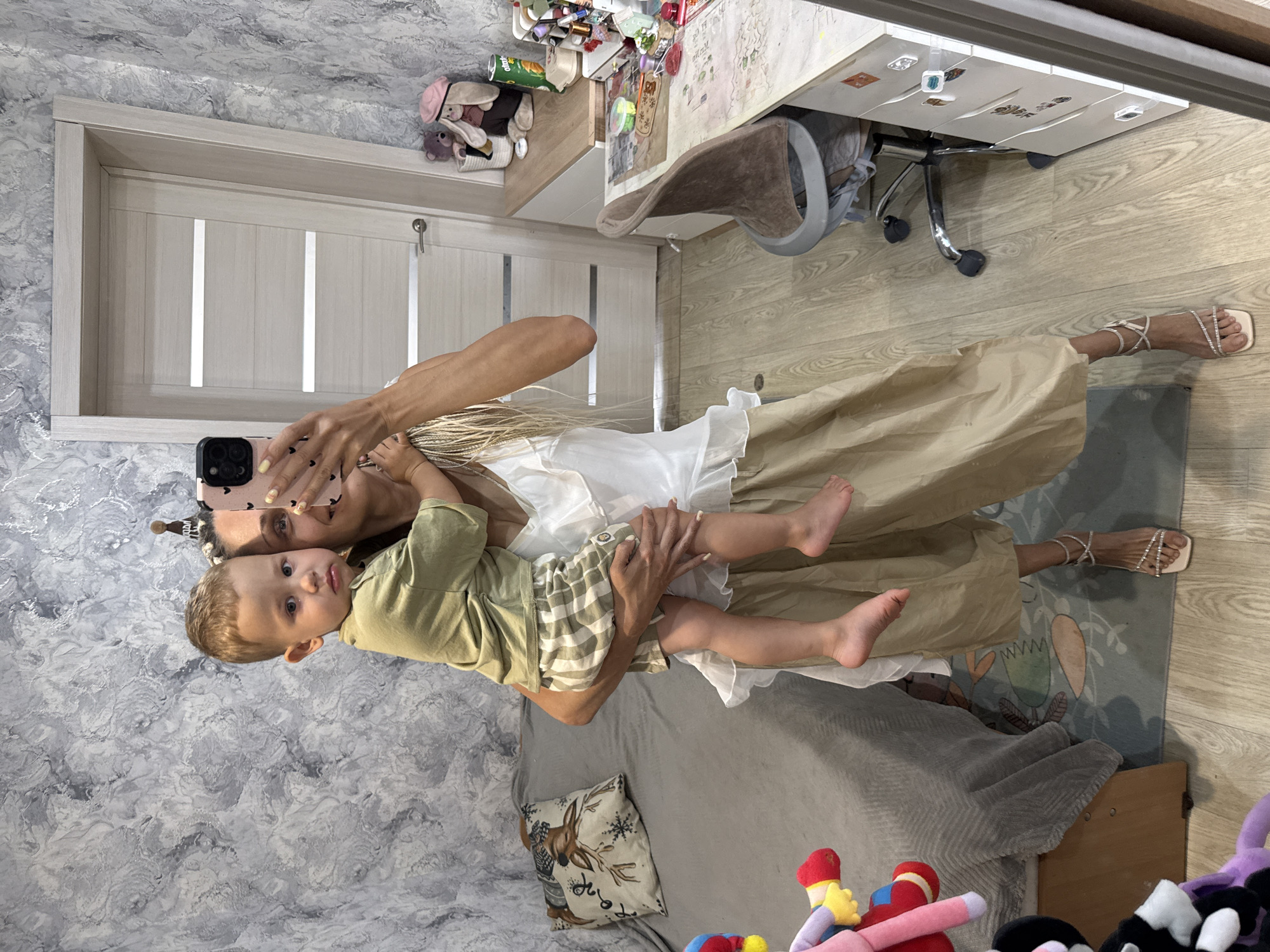 In a room with textured grey and white wallpaper, a woman is taking a mirror selfie while holding a young child. The woman has blonde hair in braids and is wearing a white ruffled top and a beige skirt. The child, wearing a green t-shirt and checkered shorts, looks at the camera with a slight frown. To the left, a grey bed with a decorative pillow depicting a deer is visible. To the right, a wooden desk is cluttered with various items, including stuffed animals, stationery, and a can of soda. A desk chair is positioned in front of the desk. A white door with horizontal panels is in the background. The floor is wooden. The location is identified as Rusanovo, Russia.