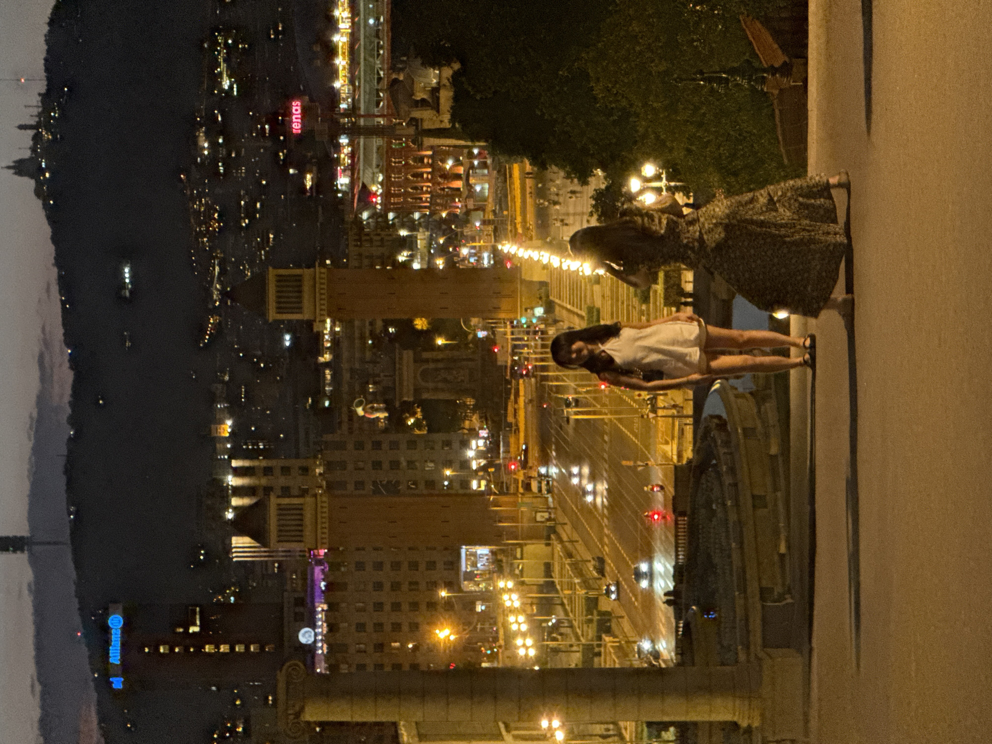 This is a night-time, medium-shot photograph of the Magic Fountain of Montjuïc in Barcelona, Spain, taken from an elevated position. The scene is dominated by the illuminated city spread across a hillside in the background, with the sky showing the twilight hours. In the foreground, two women are standing on a paved area. The woman on the left is facing the camera, wearing a white dress and black sandals. The woman on the right, facing away from the camera, is wearing a patterned dress and is holding up a phone, seemingly taking a picture. The Magic Fountain area features a semicircular pool, with illuminated stairs and a fountain visible further back, flanked by ornate lampposts. The surrounding architecture includes two prominent brick towers and several buildings lit up with artificial lights. The street below is busy with traffic, with cars and motorcycles visible, their headlights and taillights creating streaks of light. The overall atmosphere is one of urban nightlife, with the city lights creating a vibrant backdrop.