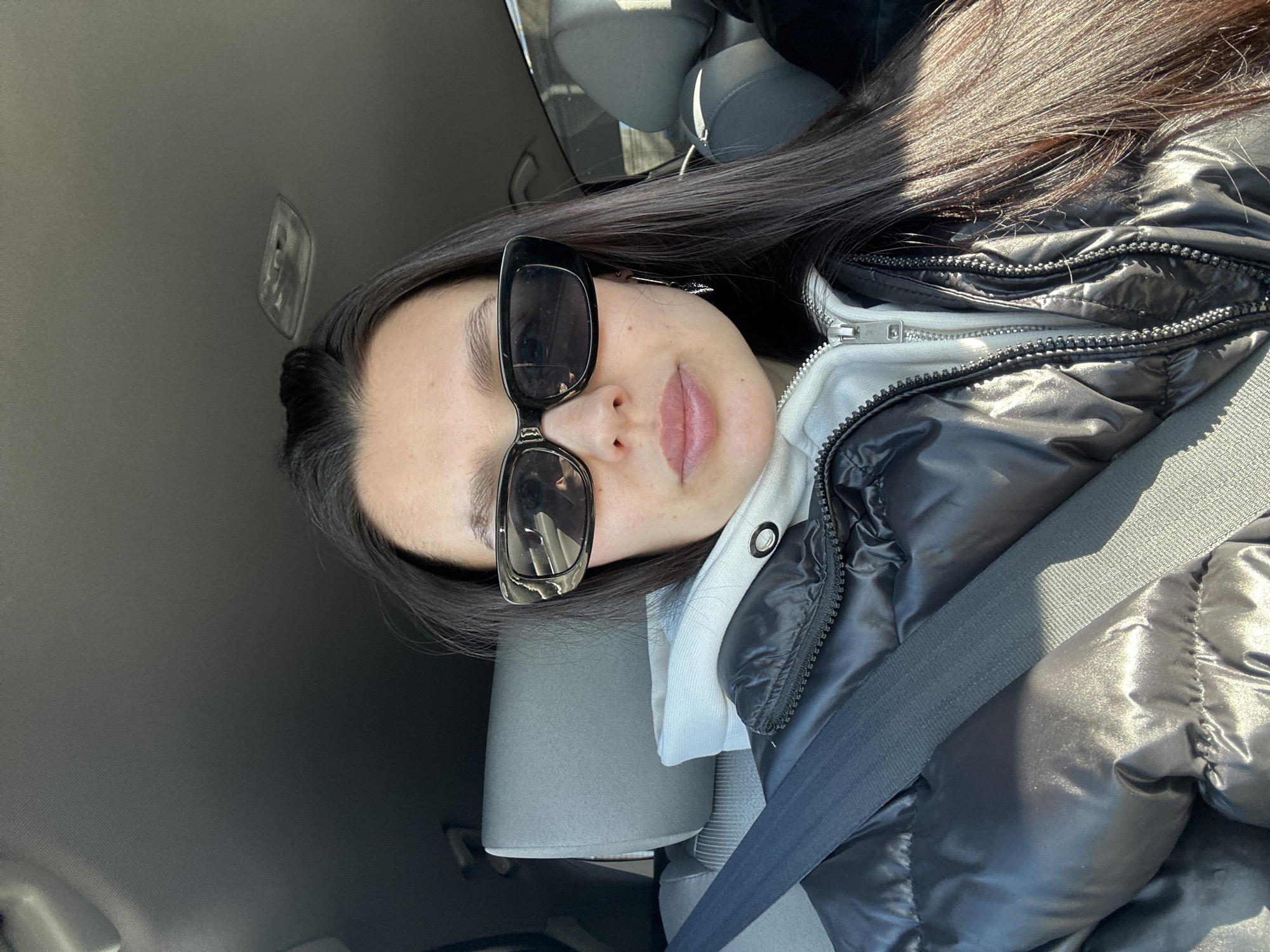 A woman with long, dark hair and sunglasses is wearing a black puffer jacket and a white hoodie while sitting in a car. She has a seatbelt fastened across her body. The interior of the car is visible, including the seats and the ceiling. The location is not identifiable as Chaikovsky, Russia, based on the image.