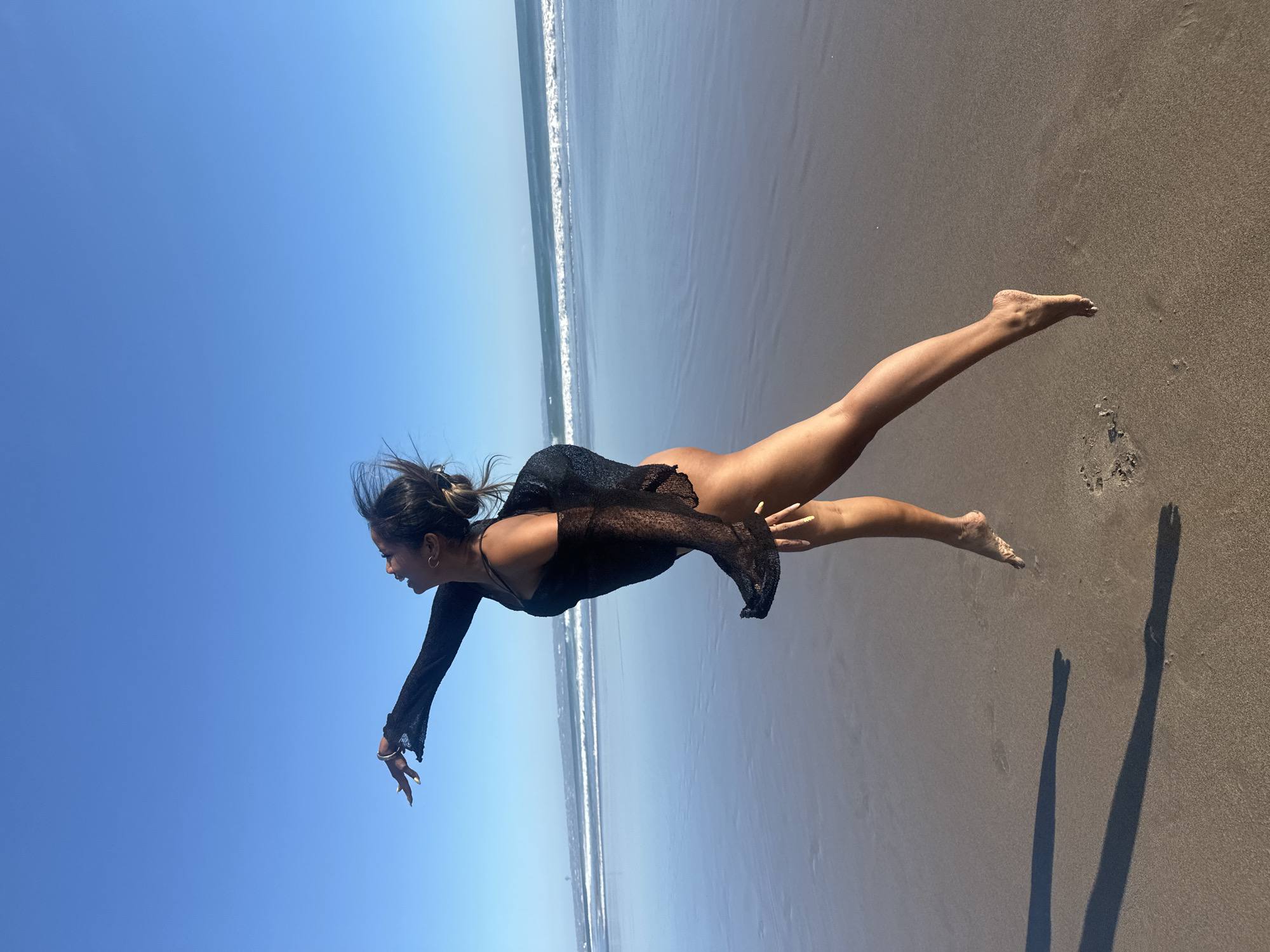 A person is captured in mid-air, appearing to float or jump across a sandy beach. They are wearing a black, sheer, long-sleeved garment over what appears to be a swimsuit. The person's legs are extended, and their arms are also outstretched, contributing to the sense of motion. The sand is a light brown, with footprints and shadows visible, indicating a sunny day. The ocean is to the left of the frame, with gentle waves breaking on the shore. The sky is a clear, vibrant blue, suggesting good weather. The lighting and long shadows indicate it is likely late afternoon or early morning. The location is identified as North Kuta, Indonesia.