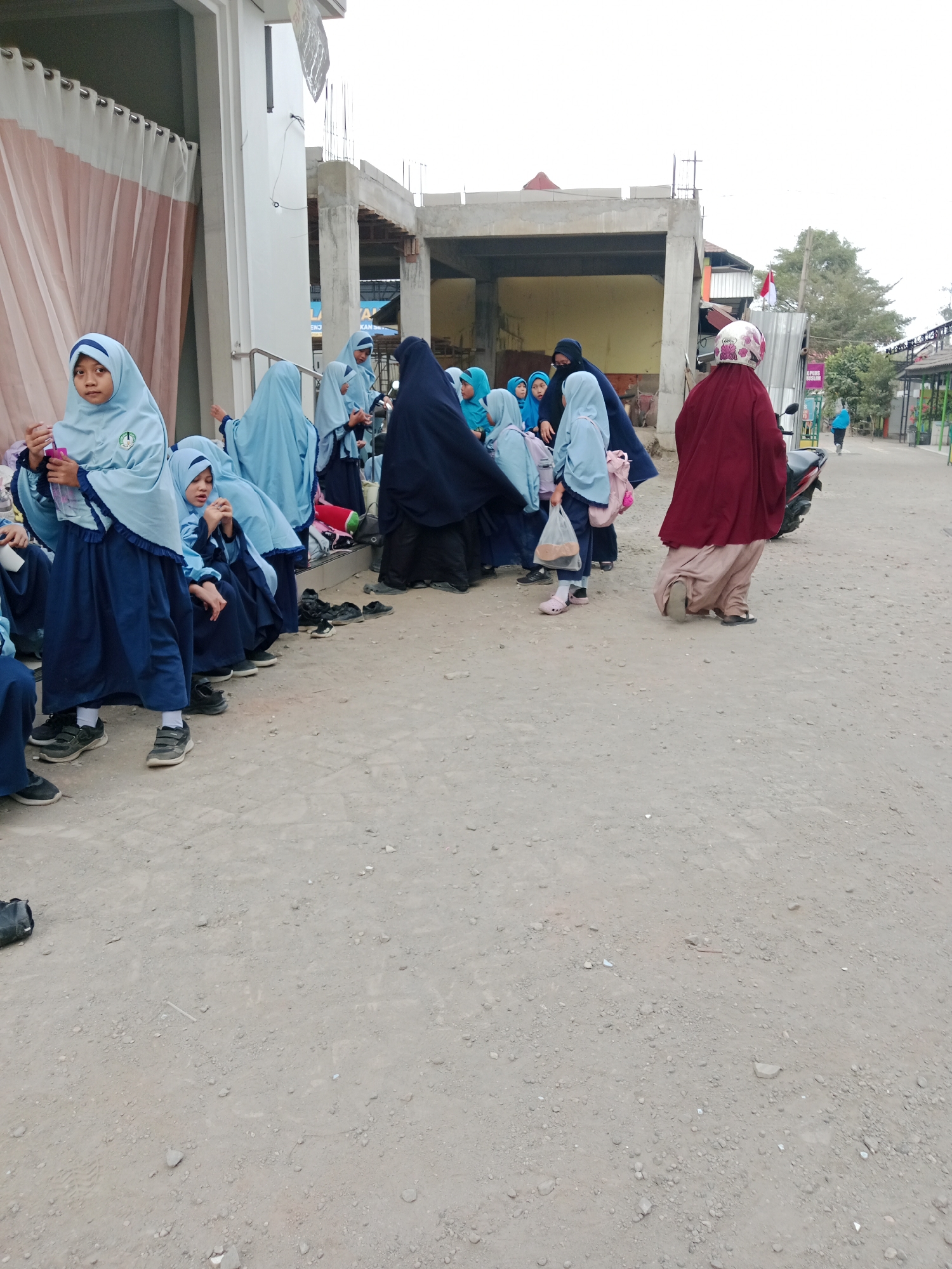 In front of a partially constructed building in Kediri City, Indonesia, a group of young girls, likely students wearing light blue hijabs and dark blue uniforms, are gathered. Some are sitting on a low step or ledge, while others stand. One girl in the foreground is holding a pink cup. Behind them, a woman in a maroon hijab and skirt is on a motorcycle, and another person in a light blue hijab is walking away in the distance. The ground is rough and dusty, with scattered gravel and debris. The building under construction has visible concrete pillars and framing. The overall scene suggests a school or a community gathering place.