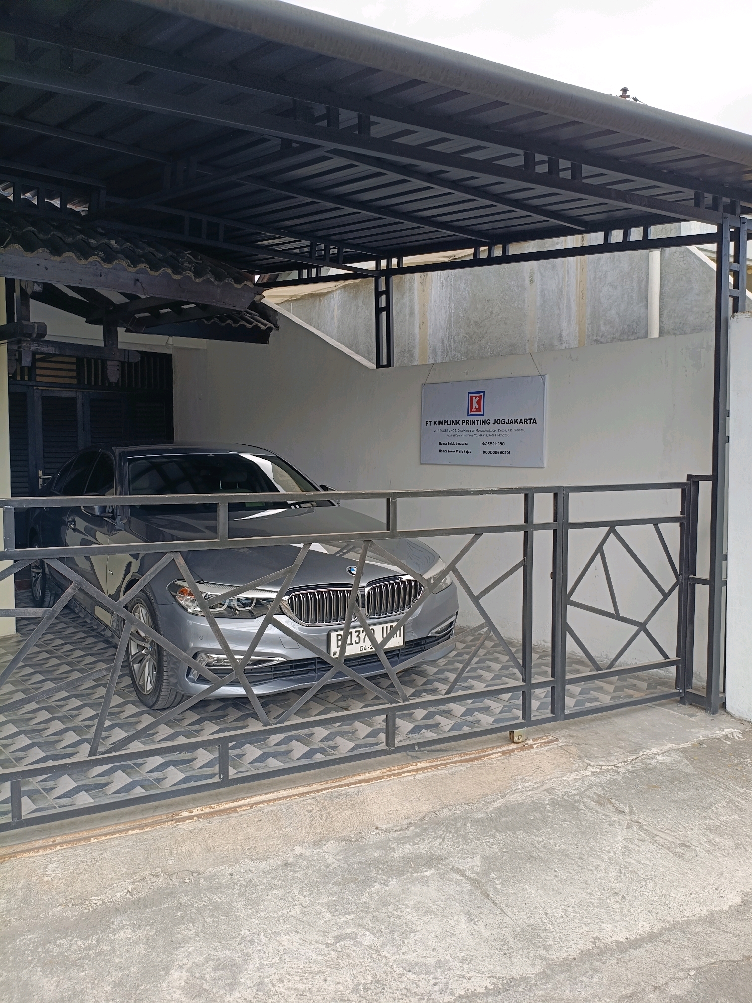 The image shows a grey BMW sedan parked under a covered carport. The carport is constructed with a dark metal frame and a corrugated metal roof. The car is positioned on a patterned tiled floor. In the background, a white wall displays a sign with the text "FT KIMPILINK PRINTING JOGJAKARTA". The sign also includes an address and phone numbers. A black metal fence with a geometric design is in the foreground, partially obscuring the car and the sign. The environment appears to be an urban setting, likely a commercial or residential area in Maguwoharjo, Indonesia, given the sign's prominent display. There are no people visible in the image.