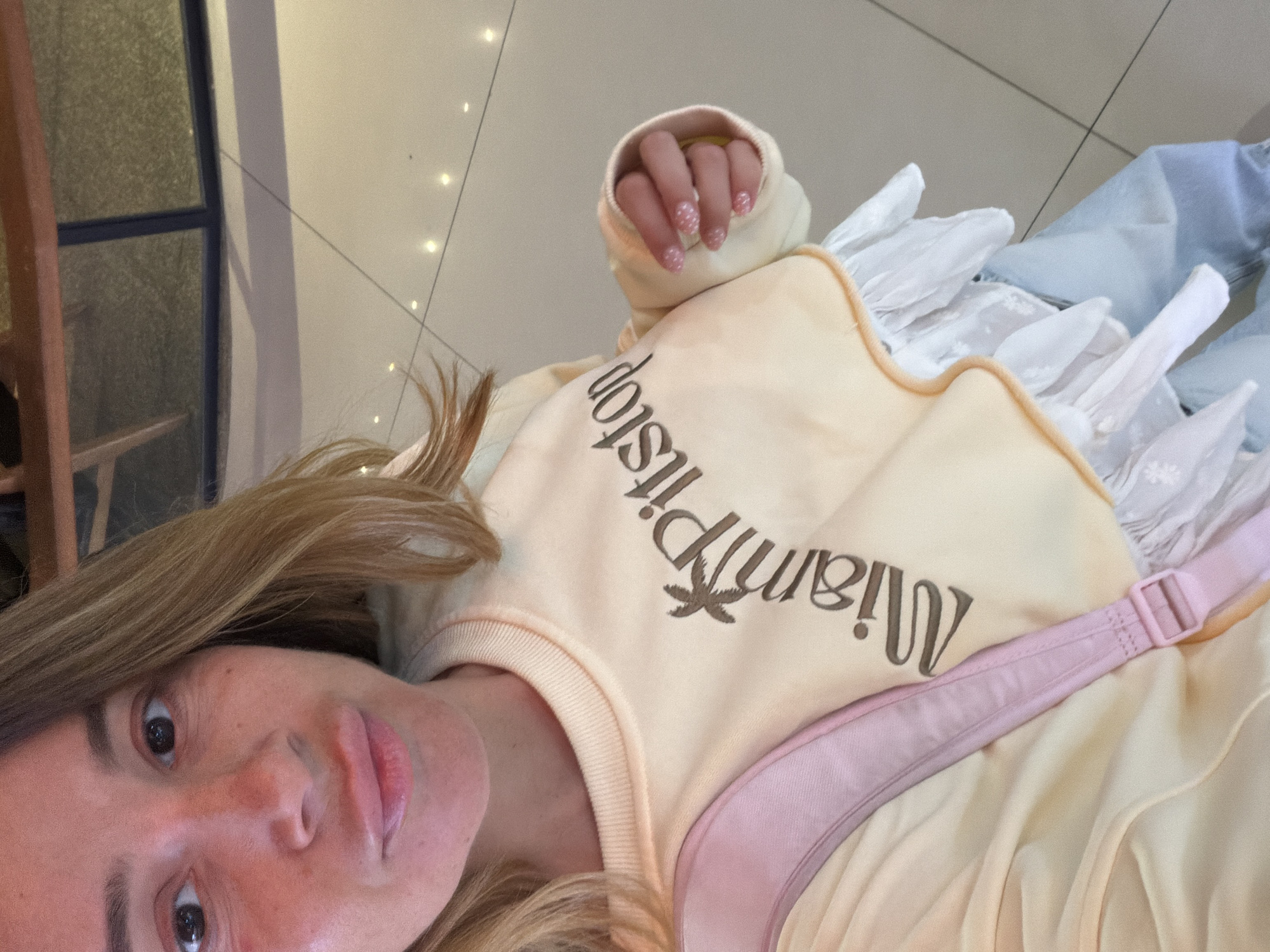 A woman, viewed from the chest up, is taking a selfie in what appears to be a public indoor space with tiled flooring. She has blonde hair and is wearing a light yellow sweatshirt with a ruffled hem and "Miami Pitstop" written across the chest in brown lettering. A pink strap is visible on her left shoulder, suggesting she is wearing a backpack. Her right hand, with decorated nails, is resting on the sweatshirt. The background is out of focus, showing a tiled floor and a reflective surface that may be a window or a glass partition. Based on the distinctive tiles and reflective surfaces often found in commercial areas, this could be a location within Kuta, Indonesia, such as a shopping mall or a hotel lobby.