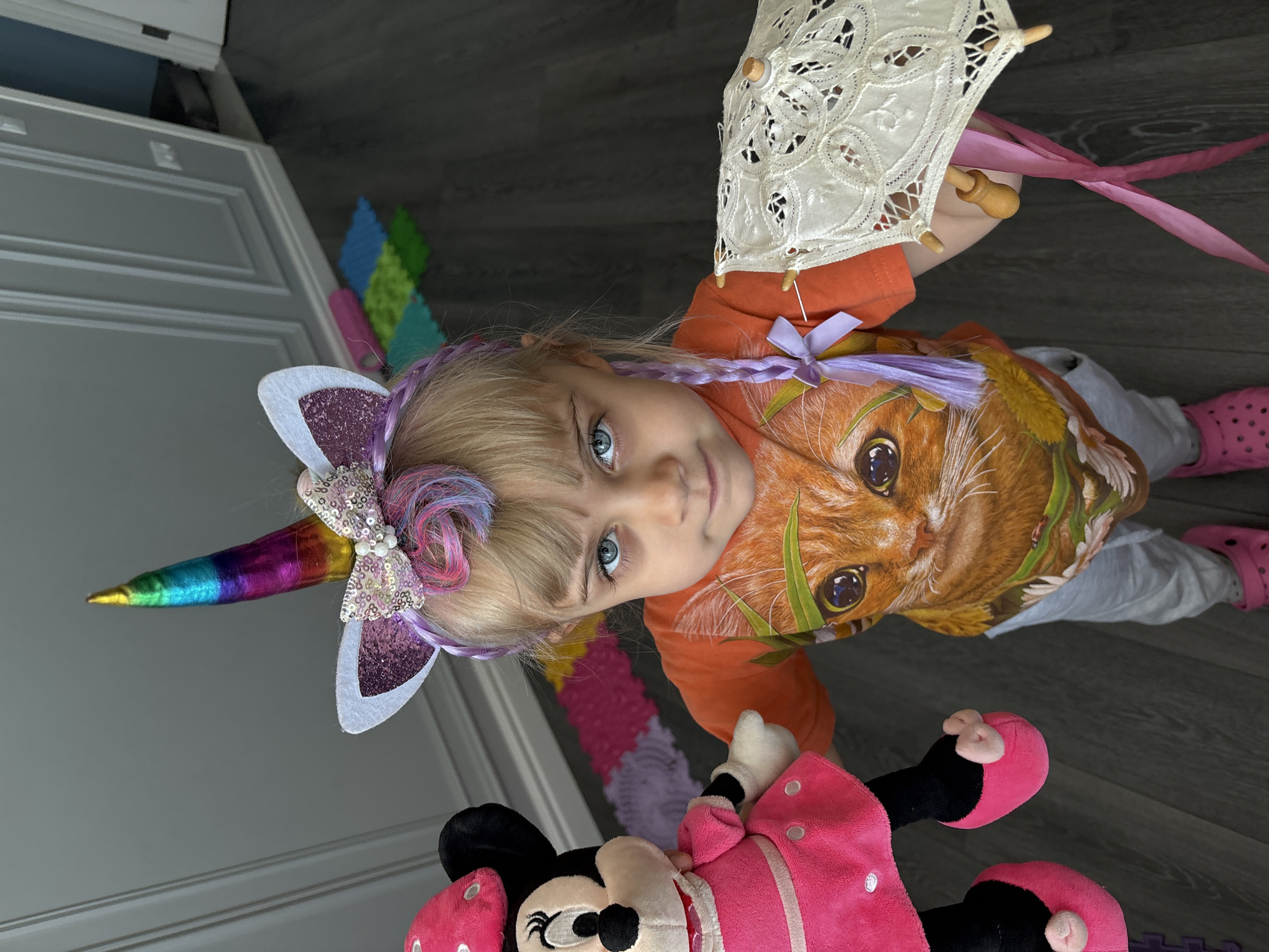 A young girl with blonde hair, blue eyes, and a unicorn headband stands on a dark gray wood floor. She is wearing an orange t-shirt with a large cat face design, gray pants, and pink Crocs. The girl is holding a pink Minnie Mouse stuffed toy in her left arm and a small white lace parasol with wooden handles and pink ribbon in her right hand. In the background, a wall with decorative molding is visible, along with several colorful puzzle mats on the floor and a partially open doorway. The lighting is bright, suggesting an indoor setting during the daytime. The overall scene appears to be a child's play area.