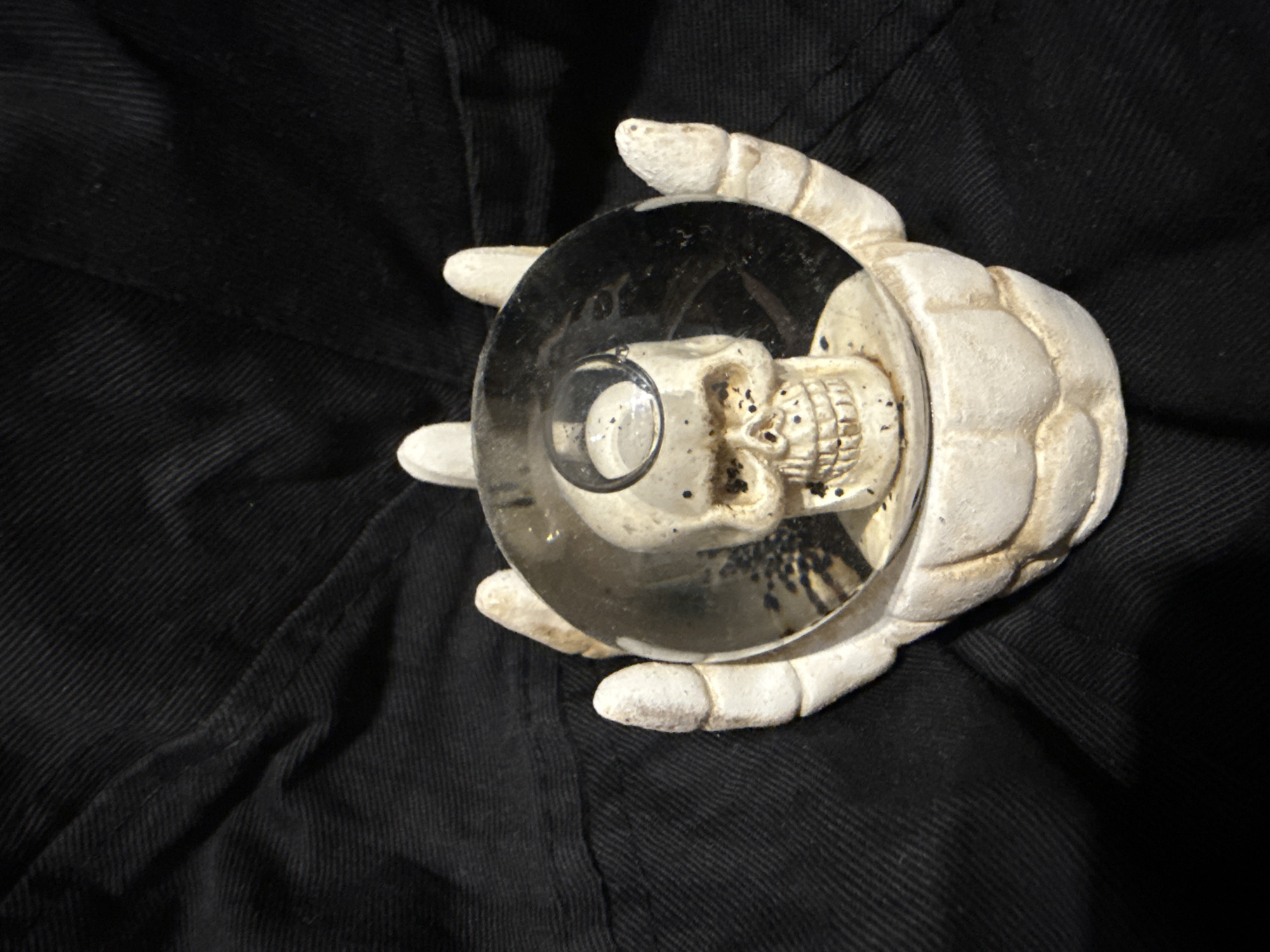 The image shows a close-up of a decorative item shaped like a skeleton's hand holding a glass orb with a skull inside. The orb is reflective and shows a distorted image of the skull. The hand and skull are off-white in color and appear to be made of resin or a similar material. The background is black fabric with visible texture. There is no discernible connection to Opoczno, Poland.