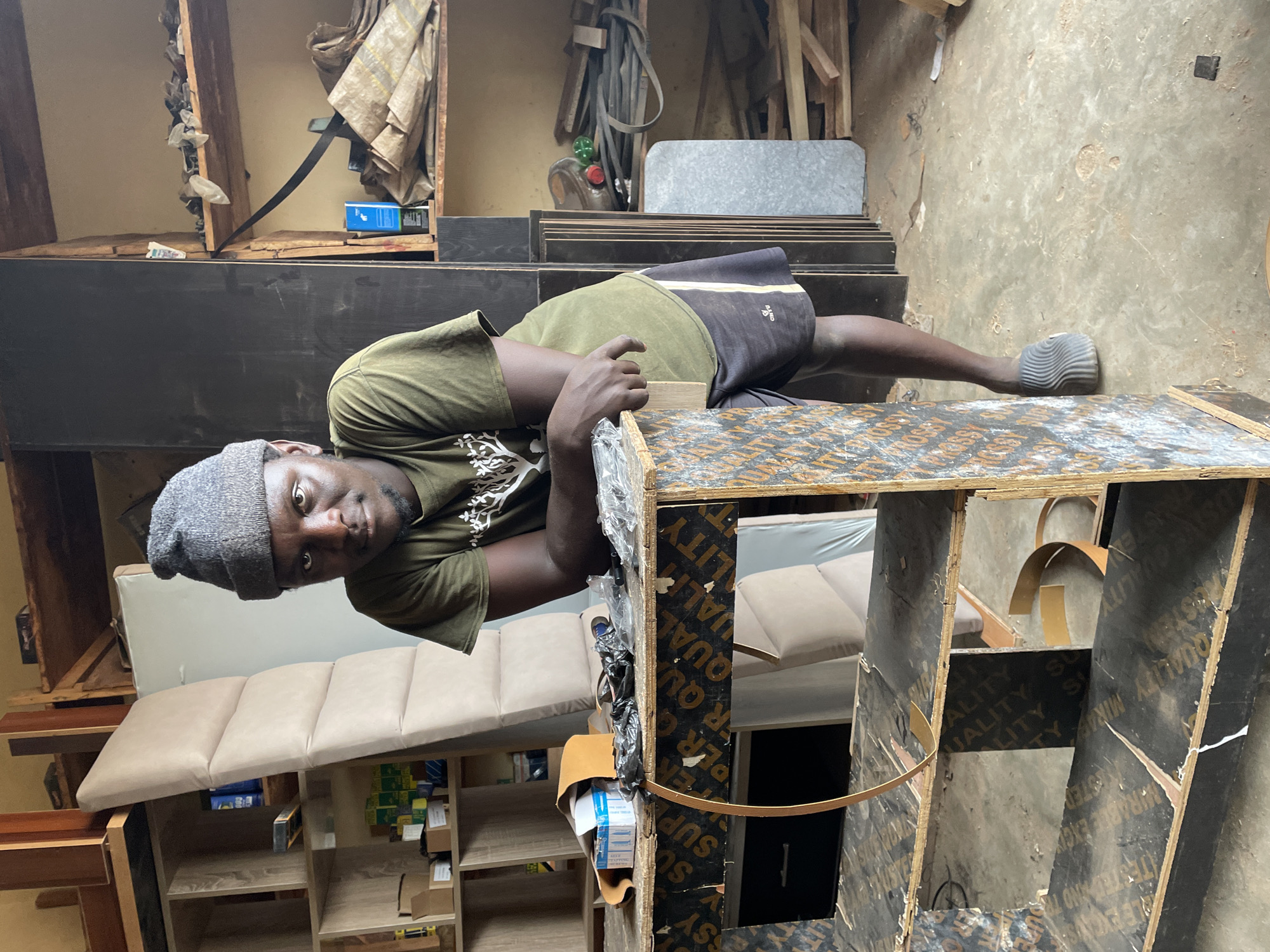 This image depicts an indoor workshop in Jos, Nigeria, likely for carpentry or upholstery. A young adult male stands centrally, leaning with both forearms on a makeshift plywood workbench, his hands clasped. He wears a grey knit beanie, an olive green t-shirt with a white tree design, dark shorts with a yellow stripe, and grey slides. He looks directly at the viewer with a neutral expression.
The workbench, constructed from dark-patterned plywood featuring "SUPER QUALITY" and "PLYWOOD" text, holds a black plastic bag, a small blue box, a brown strip of material, and other miscellaneous items. A thin, light-colored flexible strip curves over the workbench's front edge. A dark drawer unit is partially visible beneath.
To the subject's left, a light-colored wooden shelving unit displays various boxes and a blue container. A tall, light beige upholstered panel stands upright against this shelving. To the right, two additional wooden shelving units contain bundled materials, coiled straps, tools, and wood offcuts. A dark vertical panel separates these shelving sections. The floor is dusty, light-colored concrete. The scene is illuminated by ambient light.