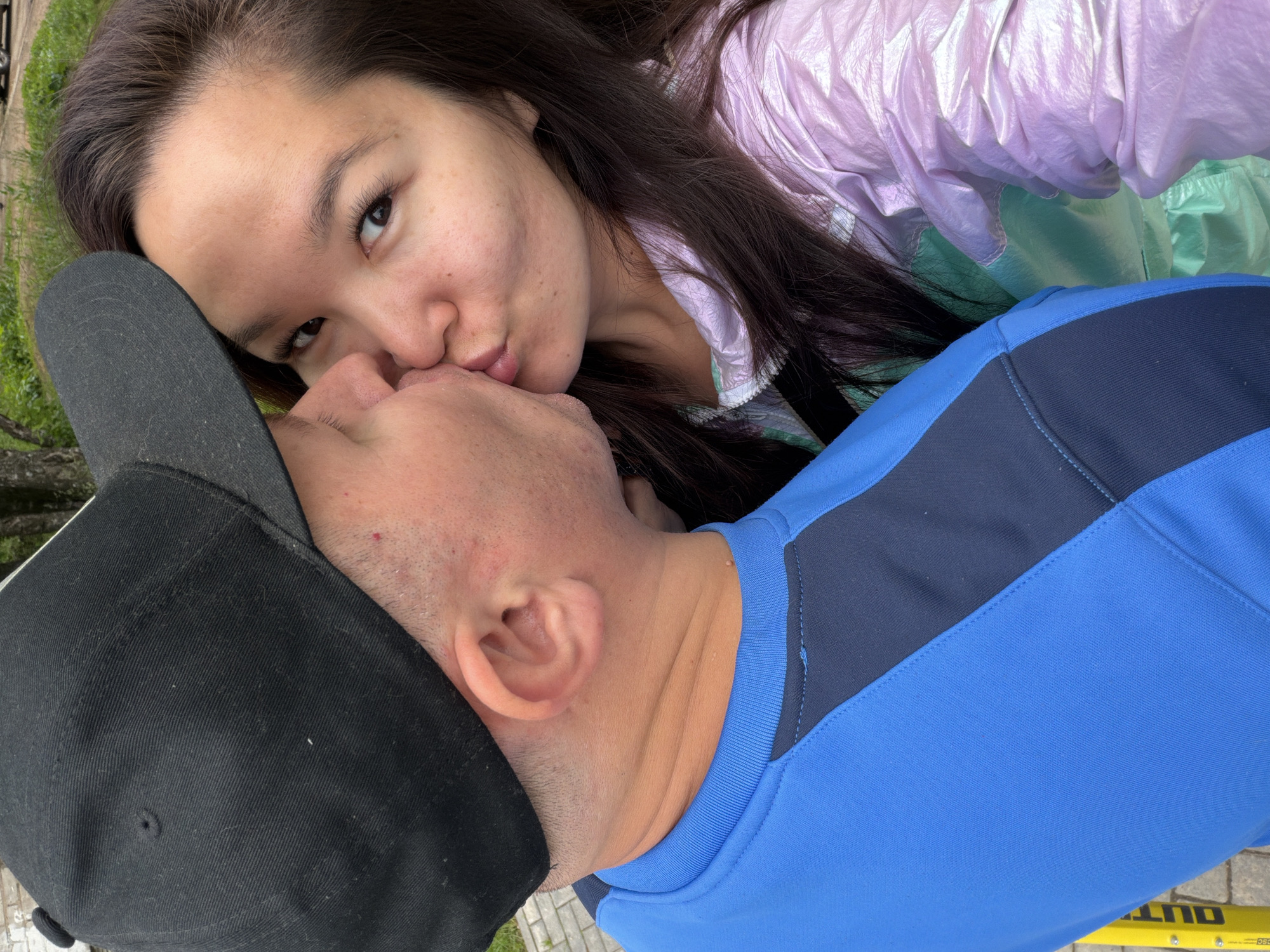 The image depicts two individuals, an adult male and an adult female, engaged in a kiss in an outdoor setting. The male is positioned on the left, partially visible from the chest up, wearing a black baseball cap and a blue long-sleeved top with a dark blue panel on the right side. His fair-toned skin shows some stubble on the cheek and jawline, and his left ear is visible. The female is on the right, also partially visible, with long dark hair. She wears a light purple iridescent jacket over a light green garment. Her face is angled towards the camera, with her eyes open and looking forward as her lips press against the male's. The background consists of green foliage in the upper section and a paved or tiled surface in the lower left, where a yellow object with the partial black text "OUT" is discernible. There are no specific landmarks or architectural features visible within the image to identify a precise location within the city of Chaikovsky, Russia.