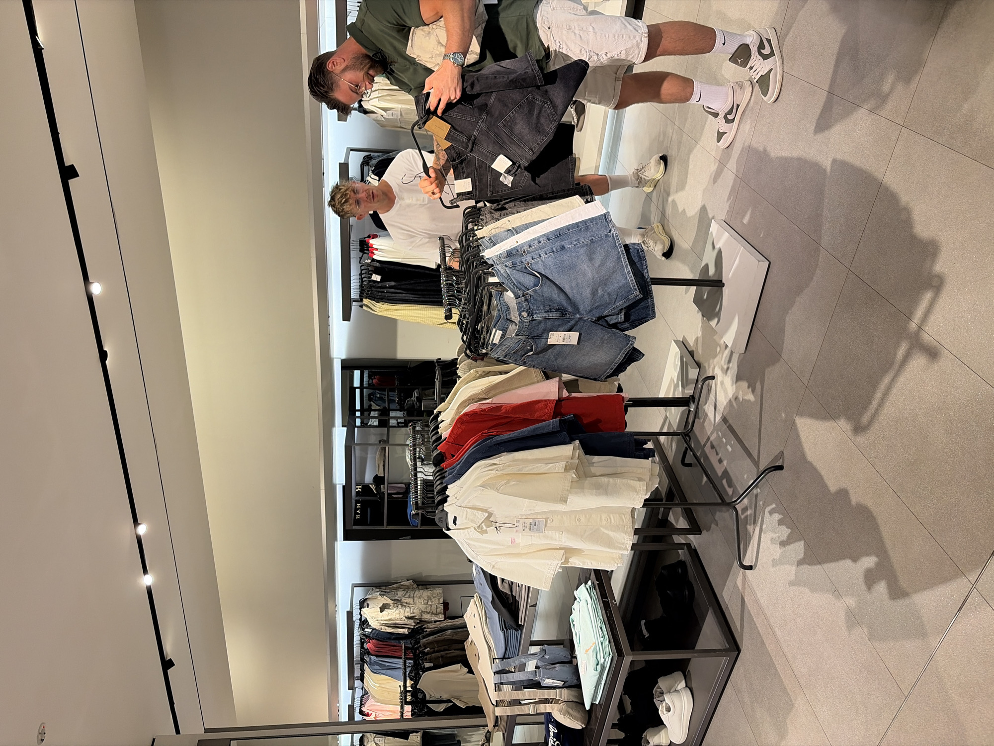Two men are browsing shorts on a clothing rack inside a brightly lit retail store, likely in Kuta, Indonesia. The man on the left, wearing a white t-shirt and glasses, holds up a pair of dark denim shorts, looking at them. The man on the right, wearing a dark green polo shirt and white shorts, examines a folded piece of light-colored fabric. A variety of men's clothing, including shirts and shorts, are displayed on racks and shelves throughout the store. The floor is tiled, and shadows from the clothing racks and people are visible. The overall scene suggests a shopping experience in a modern clothing store.