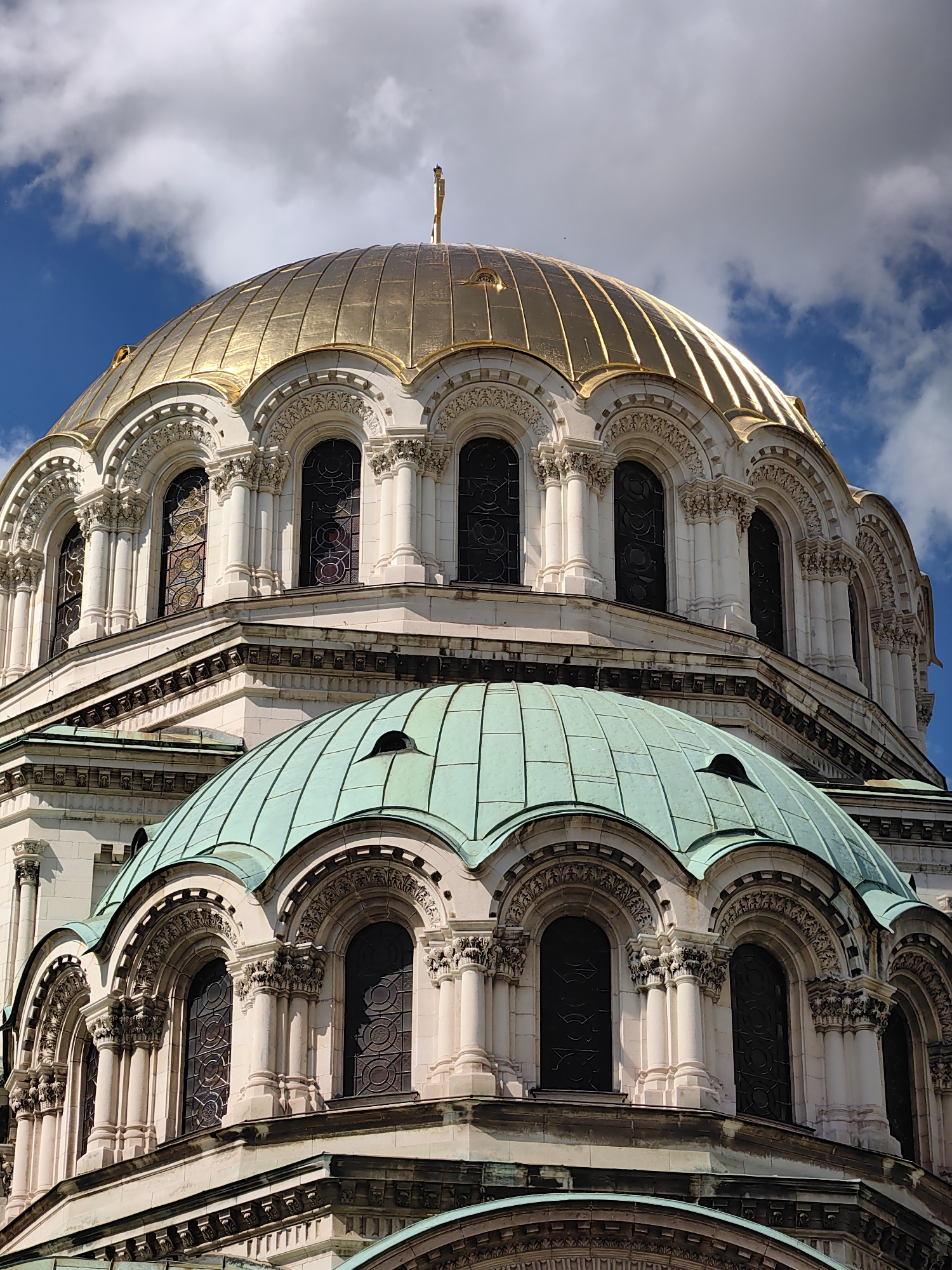The image shows a close-up view of the domes of the Alexander Nevsky Cathedral, a prominent landmark in Sofia, Bulgaria. The cathedral is an Eastern Orthodox cathedral and one of the largest in the world. The photograph focuses on the gilded central dome and a smaller, greenish-blue dome below it. Both domes feature intricate architectural details, including arched windows and decorative carvings. The sky above is a vibrant blue with scattered white clouds, suggesting a bright, sunny day. The perspective is from below, emphasizing the grandeur and scale of the cathedral's architecture. There are no people or specific actions visible in the image; it is a static architectural study.