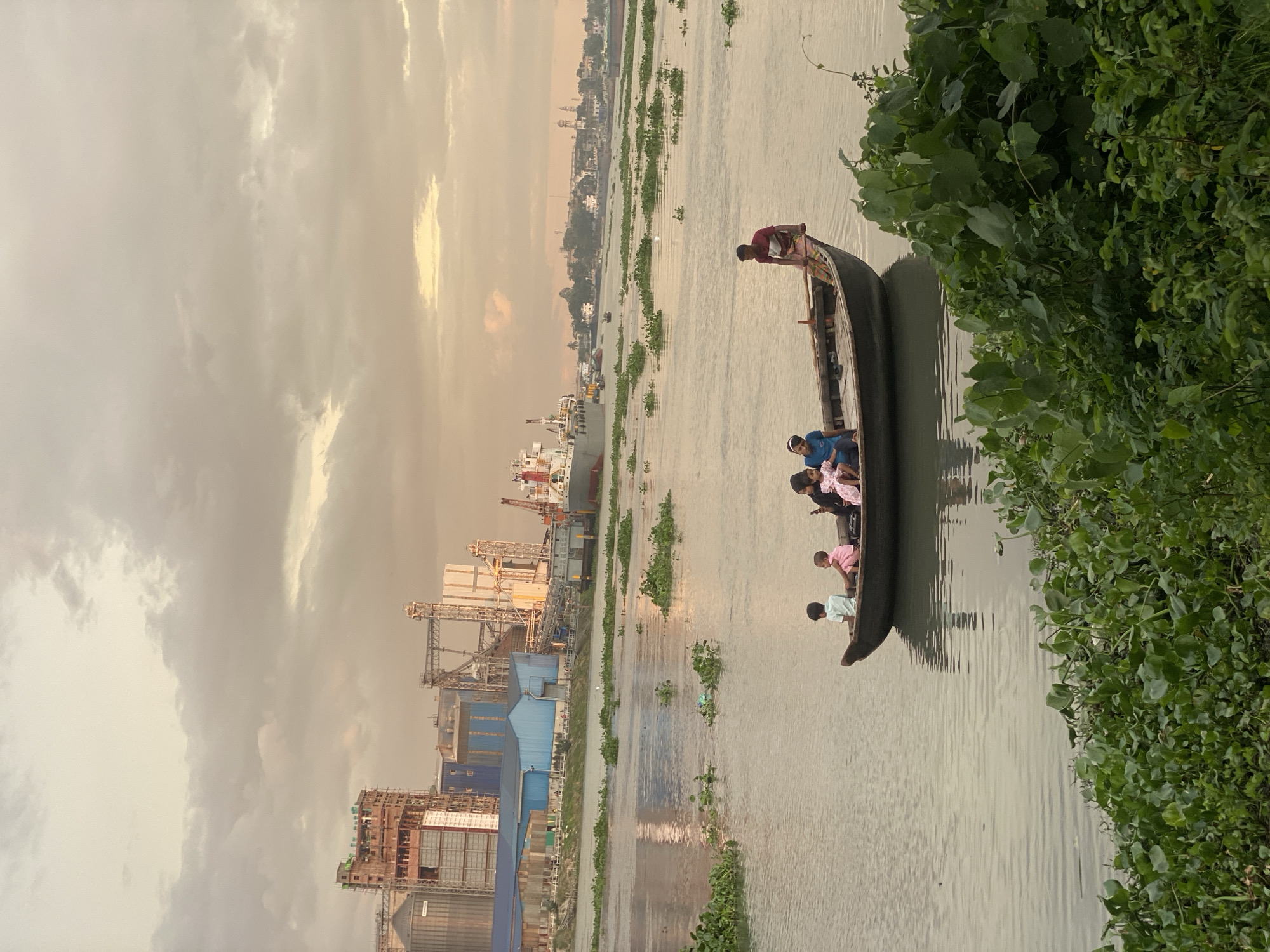 In Tarabo, Bangladesh, the sky is overcast with a mix of light and dark clouds, casting a soft, diffused light over the scene. The image captures a wide, calm river reflecting the muted tones of the sky. In the foreground, lush green aquatic plants float densely along the riverbank.
A small wooden boat, occupied by four children and an adult male, is positioned in the mid-ground. Three children are seated in the boat, while the adult male stands near the stern. The children appear to be a young boy in a light blue shirt, a younger boy in a pink shirt, and a young girl in a pink dress, with a fourth person, possibly a young girl, also seated. The adult male, wearing a red t-shirt and a patterned cloth around his waist, is looking towards the left.
In the background, a large industrial complex dominates the skyline. This complex features tall, multi-story buildings with exposed construction elements, silos, and various industrial structures. Blue and white buildings are visible, along with cranes and other machinery. Further in the distance, a large cargo ship is moored, and the outlines of more buildings suggest a populated area or another part of the industrial zone. The overall impression is one of industrial activity juxtaposed with the natural beauty of the river and its vegetation.