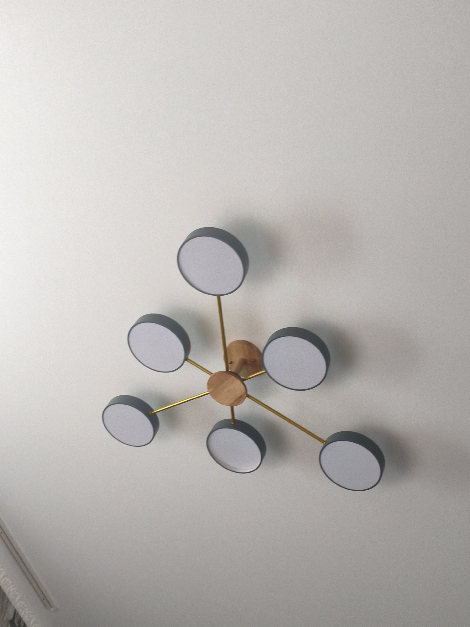 The image shows a modern, multi-arm chandelier with seven circular light fixtures, attached to a white ceiling. The chandelier has a central wooden piece from which thin, gold-colored arms extend, each holding a dark grey rimmed, white disc light. The lighting fixture is centered in the frame, casting subtle shadows on the ceiling. The context of this image within the city of Көксу, Kazakhstan is not provided by the image itself.