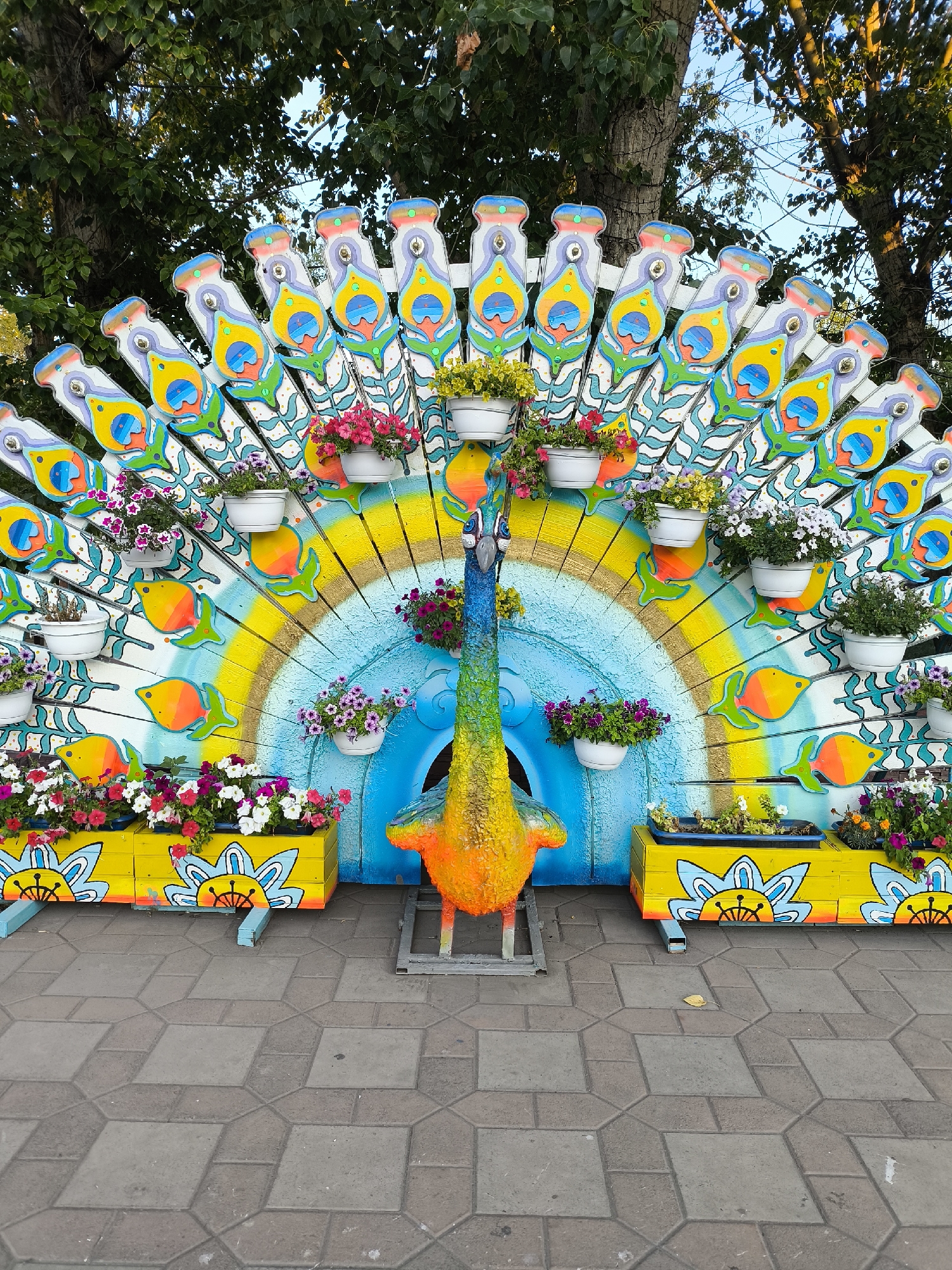 A brightly colored, decorative peacock sculpture is the central focus of this image, situated outdoors on a paved area. The peacock is fashioned from what appears to be painted wood and is adorned with many flower pots containing vibrant pink, white, and yellow flowers. The peacock's tail feathers are stylized as decorative wooden cutouts painted with peacock eye motifs, interspersed with depictions of orange fish. A circular mural in shades of blue and yellow forms the backdrop for the peacock. The sculpture is flanked by two yellow planters, also decorated with painted flower designs and holding more flowers. The ground is paved with hexagonal and square interlocking concrete tiles. Trees with green foliage and a clear sky are visible behind the sculpture, suggesting an outdoor setting, possibly a park or public space. The image is taken during daylight, with sunlight casting shadows and highlighting the vibrant colors. The location is identified as Temirtau, Kazakhstan. There are no people or animals present in the image.