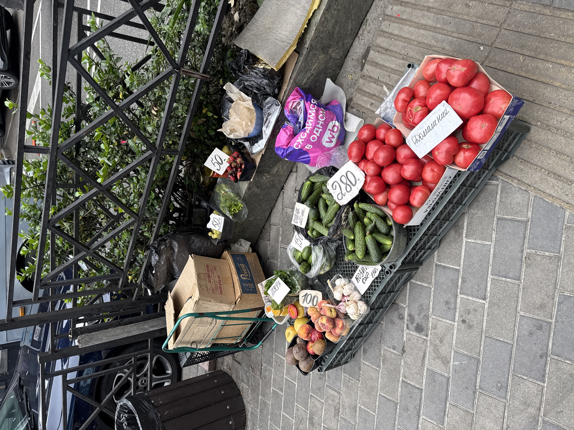 A street market stall in Sochi, Russia, displays an assortment of fresh produce, including tomatoes, cucumbers, peaches, beets, and garlic. The stall is set up on a paved sidewalk next to a metal fence and a green bush. Various plastic crates and boxes hold the fruits and vegetables, with price tags indicating "100," "150," and "280." A shopping cart with two cardboard boxes is partially visible to the left. A pink and purple reusable shopping bag with Cyrillic text and a white "wb" logo rests on the ground. In the background, a dark blue car is parked. The overall scene depicts a typical outdoor market environment in an urban setting.