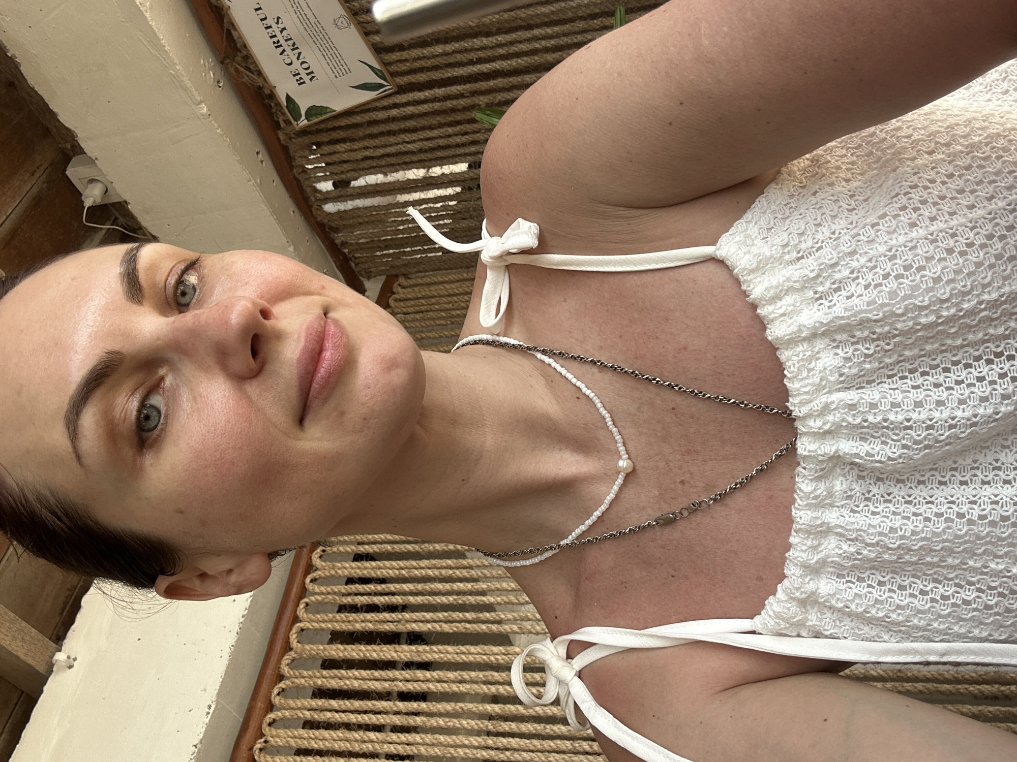 A woman with fair skin, green eyes, and dark hair pulled back is shown in a selfie taken in Kuta Selatan, Indonesia. She is wearing a white, lacy top with thin straps tied at the shoulders and two delicate necklaces, one with small white beads and a pearl, and another with a silver chain. The background includes a woven rope-like structure and a sign with text that says "BE CAREFUL MONKEYS." A white beam and electrical outlet are also visible. The scene suggests an outdoor or semi-outdoor setting.