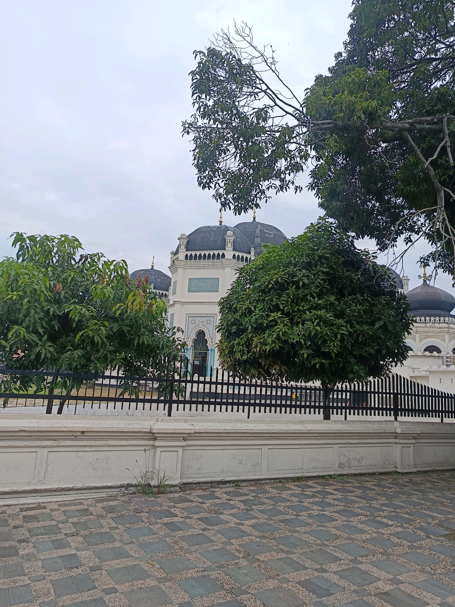 The image shows the exterior of the Great Mosque of Medan in the city of Medan, Indonesia. The mosque features prominent black domes and white architecture with intricate decorative elements. In the foreground, there is a black metal fence and a low white wall. Beyond the fence, several trees with lush green foliage partially obscure the view of the mosque. The ground in the foreground is paved with a pattern of dark and light gray square tiles interspersed with small pebbles. The sky is overcast and appears light gray. There are no people visible in the frame. The scene is captured from a low angle, looking up towards the mosque.