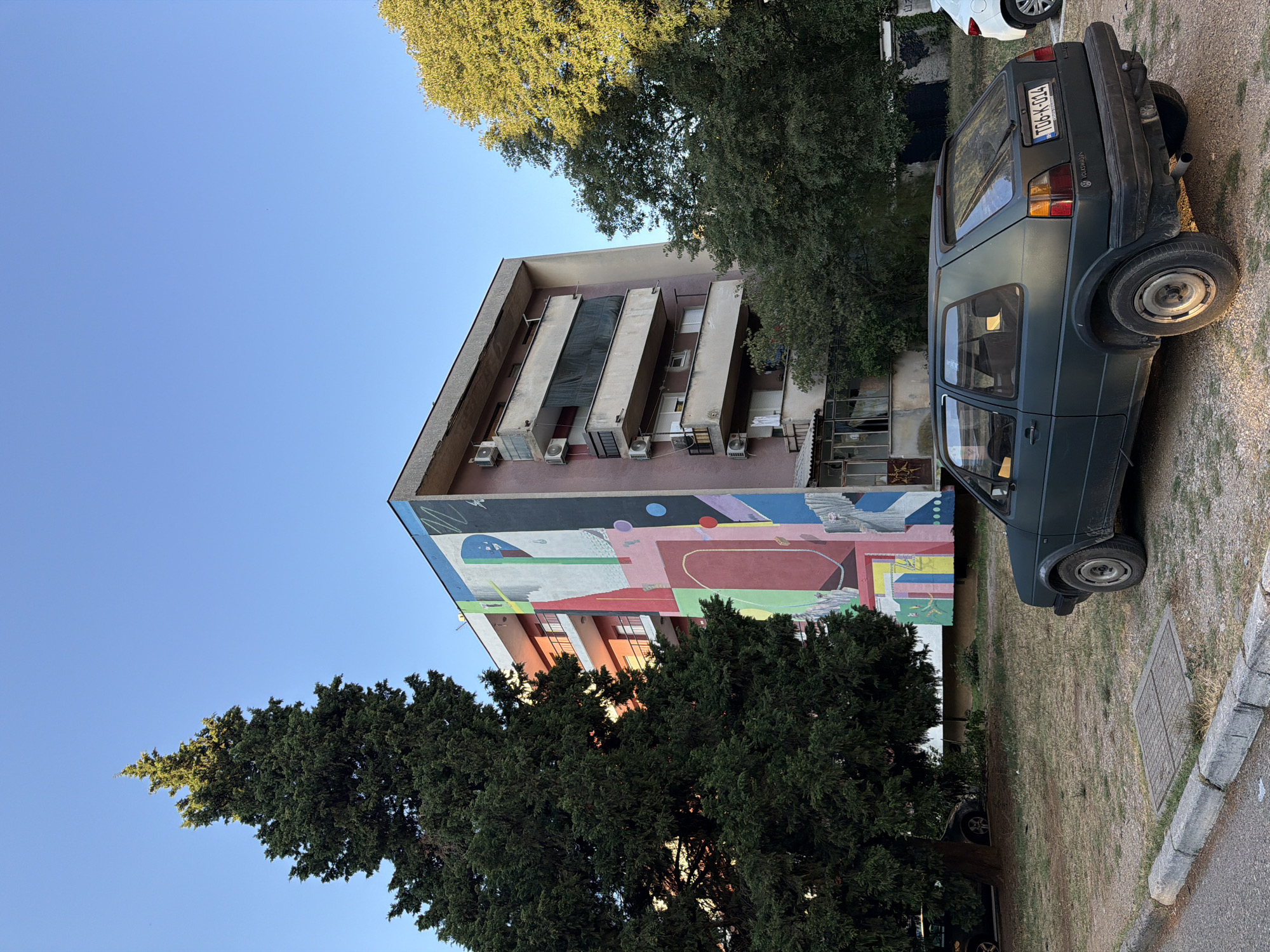 In Mostar, Bosnia and Herzegovina, a dark green Volkswagen Golf Mk2 is parked on a dirt lot next to a sidewalk. The car is facing right and its rear is visible. The license plate reads "TG6-K-014". Behind the car is a building with a large, colorful mural painted on its side. The mural features abstract geometric shapes in various colors, including red, pink, yellow, blue, and white. The building appears to be a multi-story apartment complex with balconies. A tall, dark green evergreen tree stands to the left of the building, and other trees with green foliage are visible on the right. The sky is clear and blue.