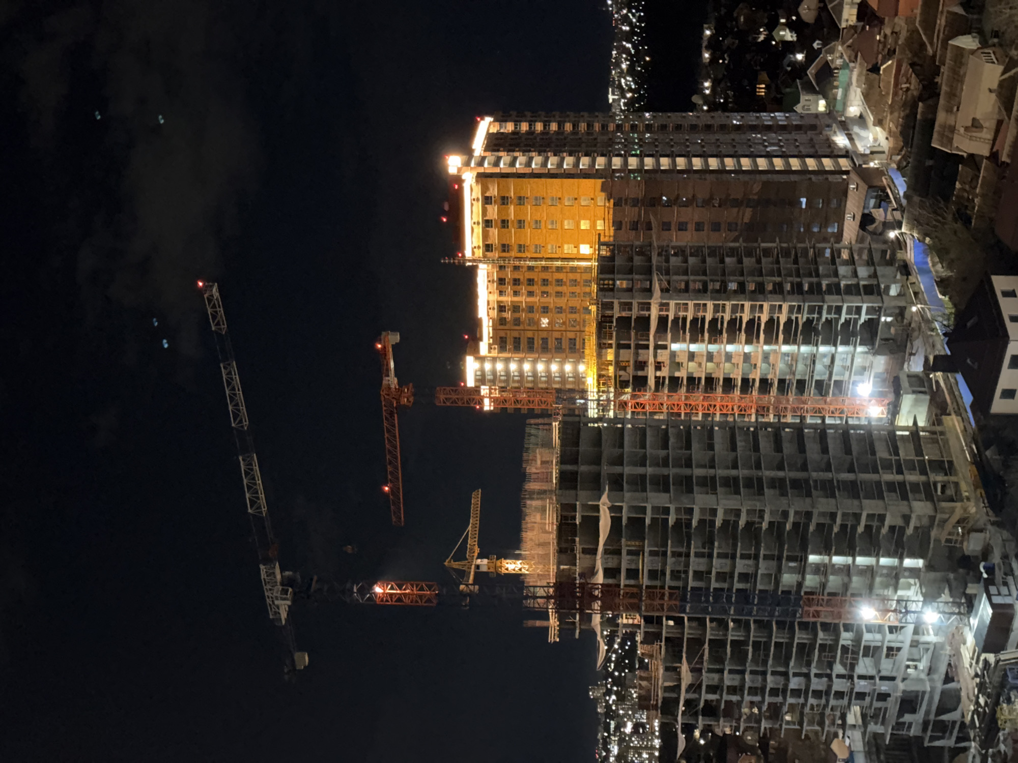 The image shows a night view of a large construction site in what is likely Rostov-on-Don, Russia. Several high-rise buildings are under construction, with varying levels of completion. The unfinished structures have exposed frameworks, illuminated by artificial lights, highlighting the skeleton of each floor. Construction cranes are visible, standing tall against the dark sky, with a few appearing to be in operation. Residential buildings and city lights surround the construction project. The lighting contrasts between the brightly lit construction site and the dimmer ambient urban environment.