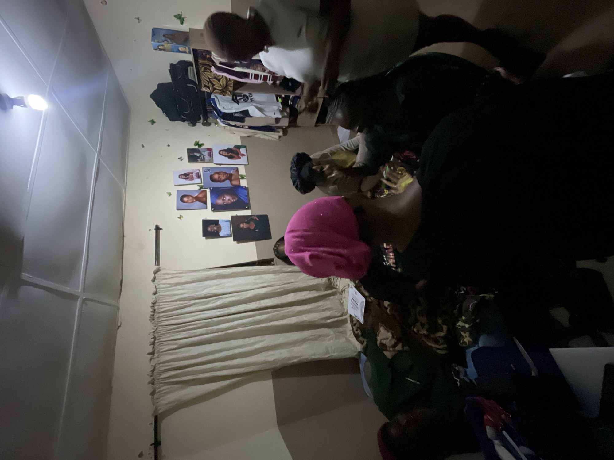 The image depicts an indoor scene, likely a room, with several individuals gathered. The room appears to be dimly lit, with a single lightbulb hanging from the ceiling providing illumination. On the wall, there are several framed photographs of people. A curtain hangs on one side of the room, and clothing items are visible on a rack in the background. The floor is covered with a patterned blanket, and some personal belongings are scattered around. The individuals are seated or standing, and their interactions are not clearly discernible due to the low light conditions and the angle of the photograph. There is no visible indication of the city of Durimi, Nigeria, within the image itself.