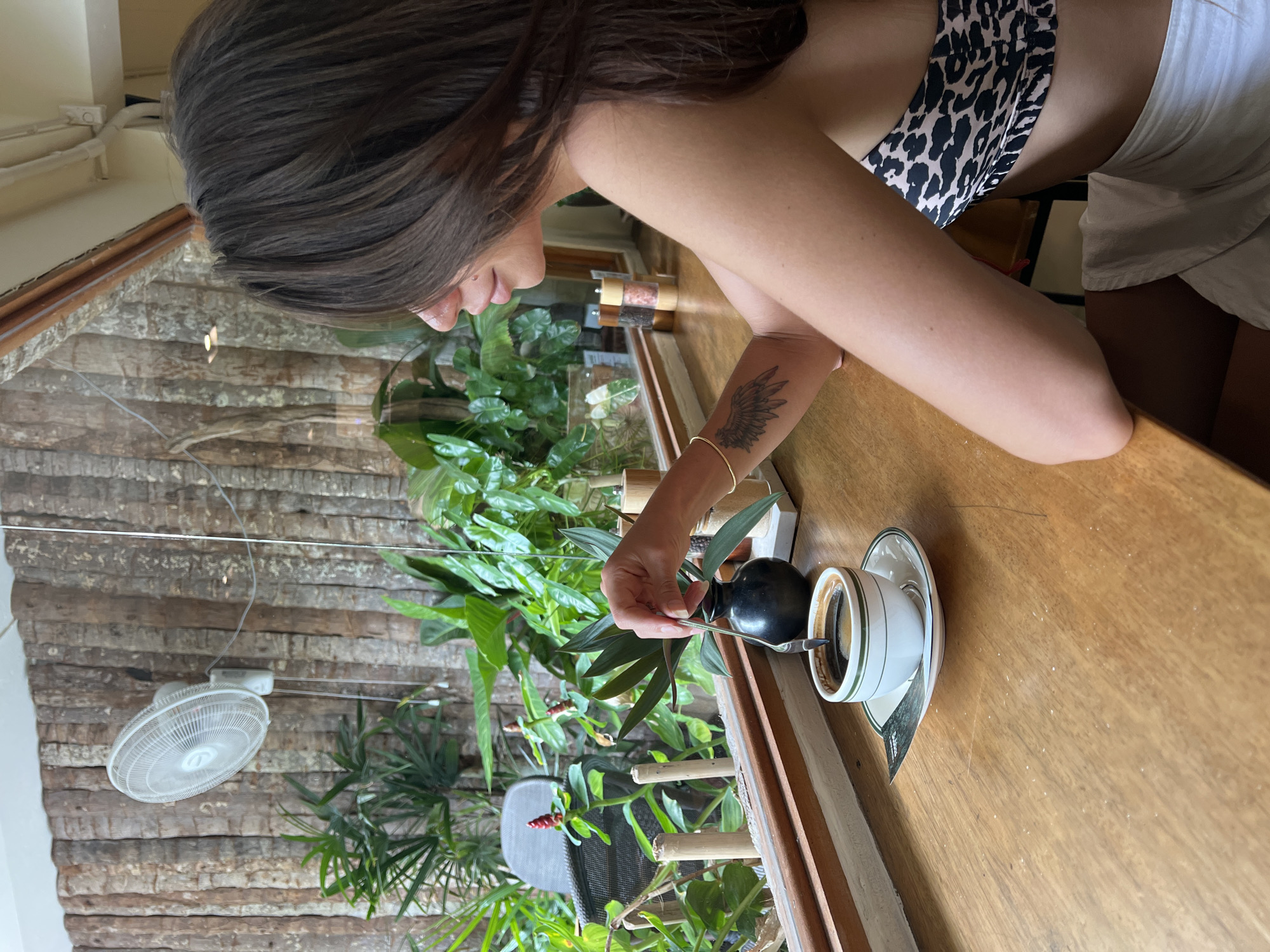 A young woman with dark hair is seated at a wooden table in front of a window. She is wearing a leopard print top and white shorts. She is holding a spoon in a white cup of coffee. Outside the window, there are palm trees, plants, and a ceiling fan mounted on a wall made of logs. The scene appears to be a cafe or restaurant in Koh Phangan, Thailand.