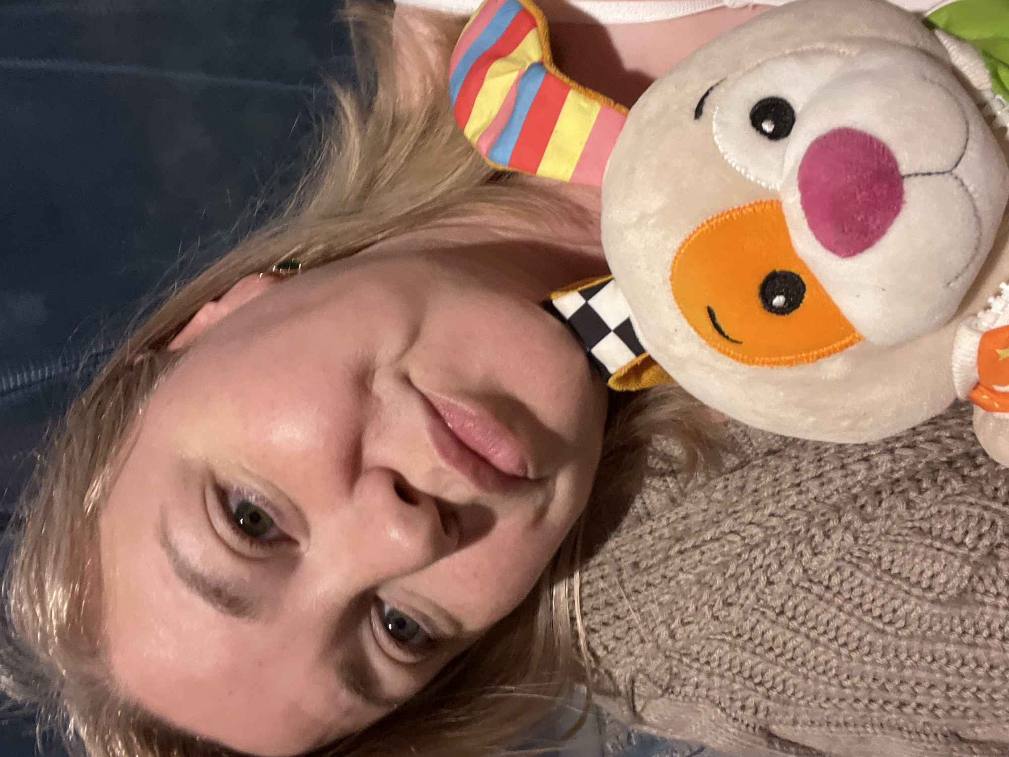 A fair-skinned woman with blonde hair is lying down, her head tilted to the side. She has light eyes and a slight smile. Next to her is a plush toy with a white head, an orange face, a pink cheek, and black eyes. The toy has a striped colorful ear, and a checkered black-and-white pattern is also visible. The woman is wearing a beige knitted sweater. The background appears to be a dark blue or gray surface. There is no verifiable evidence in the provided image to confirm it was taken in Vesyoloye, Russia.