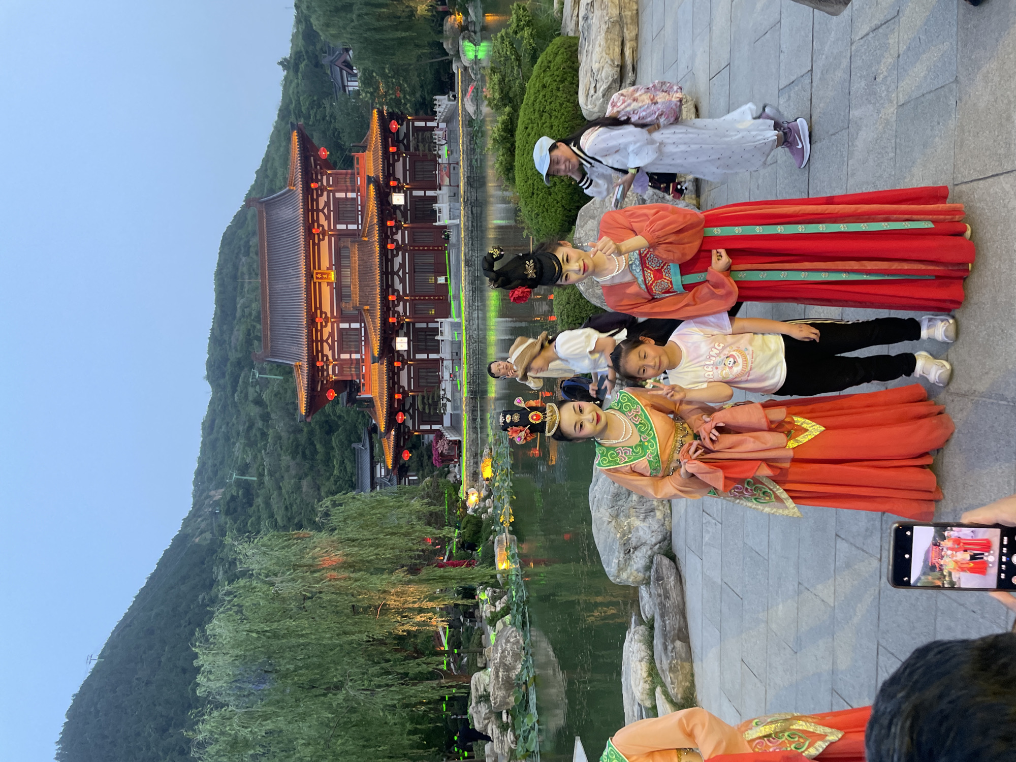 The image captures a group of people, including two women in traditional Chinese costumes, posing for a photo near a body of water in what appears to be a park or garden setting. The photo is being taken by someone using a smartphone. In the background, there is a large traditional Chinese building with red lanterns hanging, set against a green hillside. The location is Lintong District, China. The environment suggests an evening scene with the lights reflecting on the water.