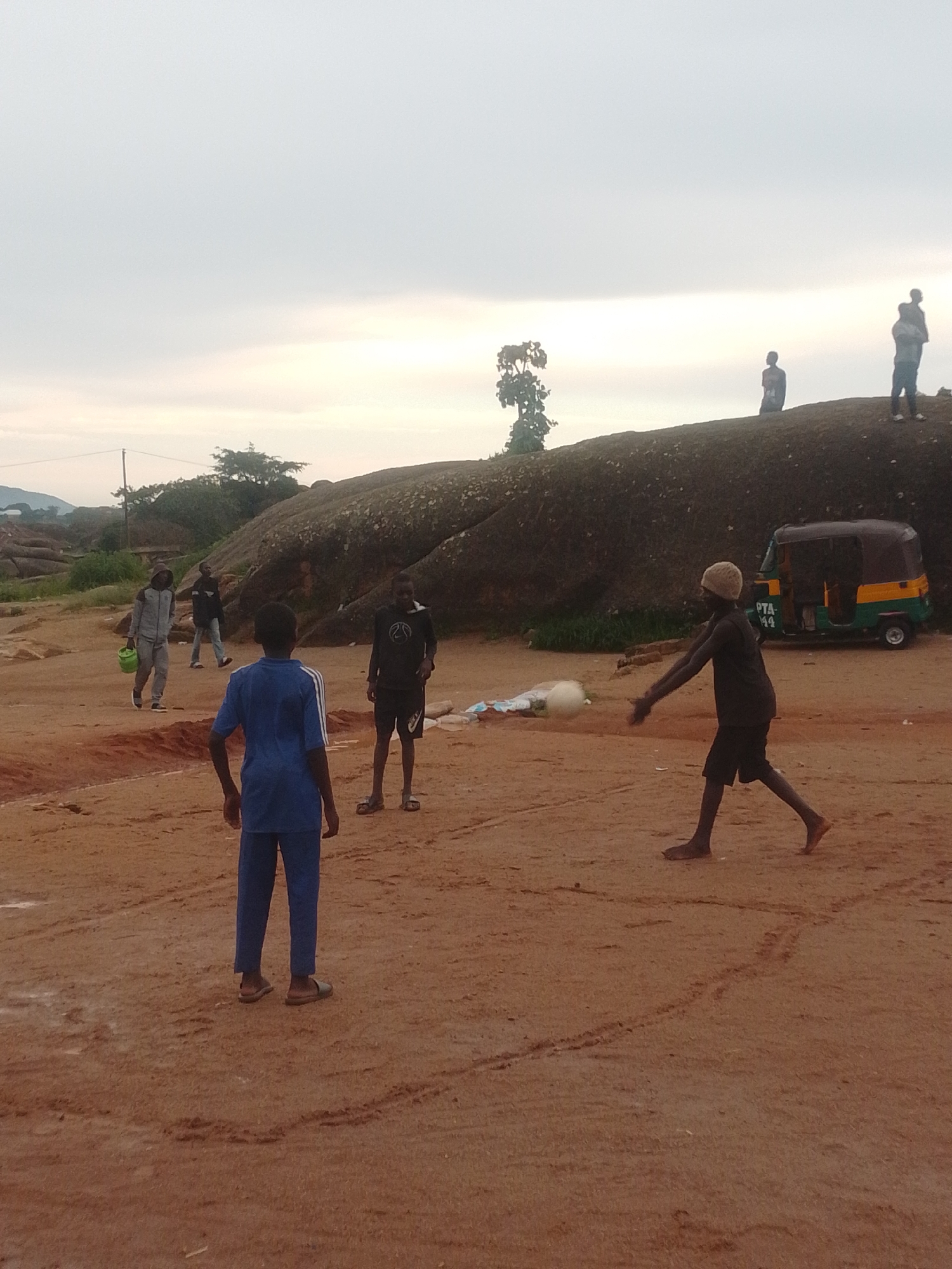 A group of young males are engaged in a game of volleyball on a sandy, reddish-brown terrain. The scene is outdoors, with a large, weathered rock formation dominating the background. The sky is overcast with a pale, diffused light, suggesting it might be dawn, dusk, or a heavily clouded day. Several figures are scattered across the foreground and mid-ground.
In the foreground, a young male in a blue t-shirt and dark blue pants stands with his back to the camera, facing the game. To his right, another young male, wearing a dark t-shirt and dark shorts, is in motion, seemingly having just served or hit a white volleyball. He is barefoot and wears a light-colored beanie. Further to the left, a young male in a black t-shirt and black shorts is standing, watching the play. Another individual, wearing a gray hooded jacket and light-colored pants, is walking towards the left, carrying a green bucket. Behind him, another person is walking away from the camera.
In the mid-ground, near the large rock formation, a three-wheeled auto-rickshaw, colored green and yellow with a license plate reading "PTA 144," is parked. There are some white plastic bags and other debris on the ground near the auto-rickshaw. The rock formation itself is massive and uneven, with vegetation growing around its base and on its slopes. In the far background, more rocky hills and trees are visible under the subdued sky. The overall impression is of a casual outdoor gathering and recreational activity in a somewhat undeveloped, natural setting.