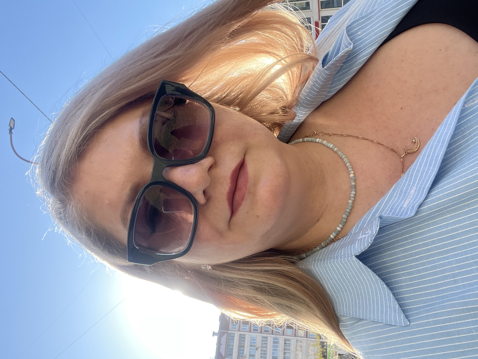 A woman is pictured in a selfie taken outdoors. She is wearing sunglasses and a light blue striped shirt. A beaded necklace and a gold necklace with a moon pendant are visible around her neck. Part of a building is seen in the background, along with a lamp post and a clear blue sky. The location is likely Izhevsk, Russia, based on the building architecture.