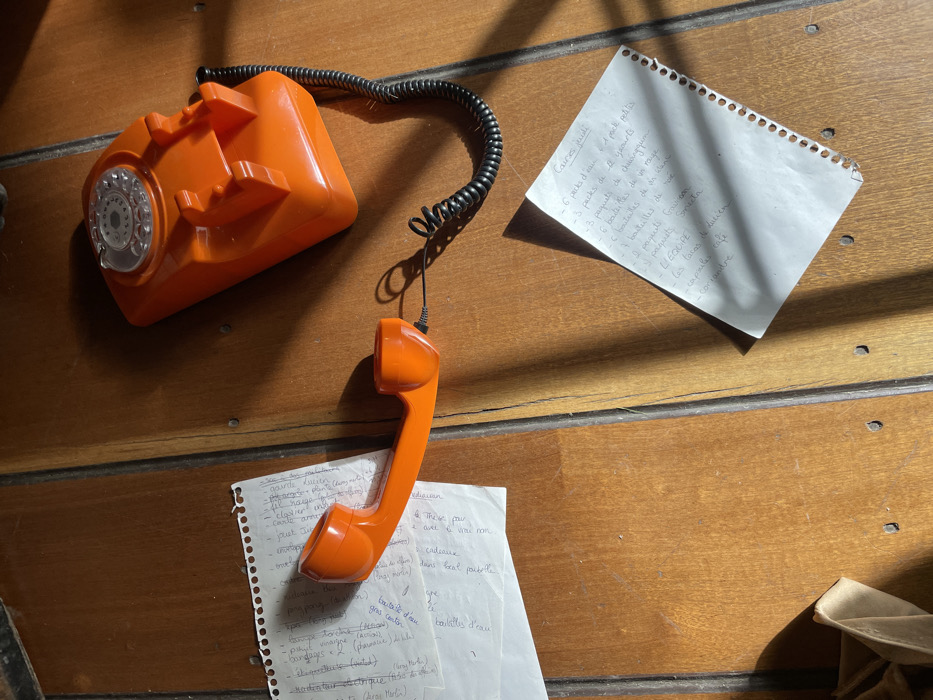 An orange rotary phone sits on a wooden surface, with its receiver dangling. Two pieces of paper with handwritten notes are also present. One appears to be a shopping list, including items like water, yogurt, chewing gum, wine, and medication such as Gaviscon and Smecta. The other list seems to have items such as "gourde Lucien", "cadeaux", "lampe torche", and "rodenticide électrique". Shadows cast by a light source add depth to the scene.