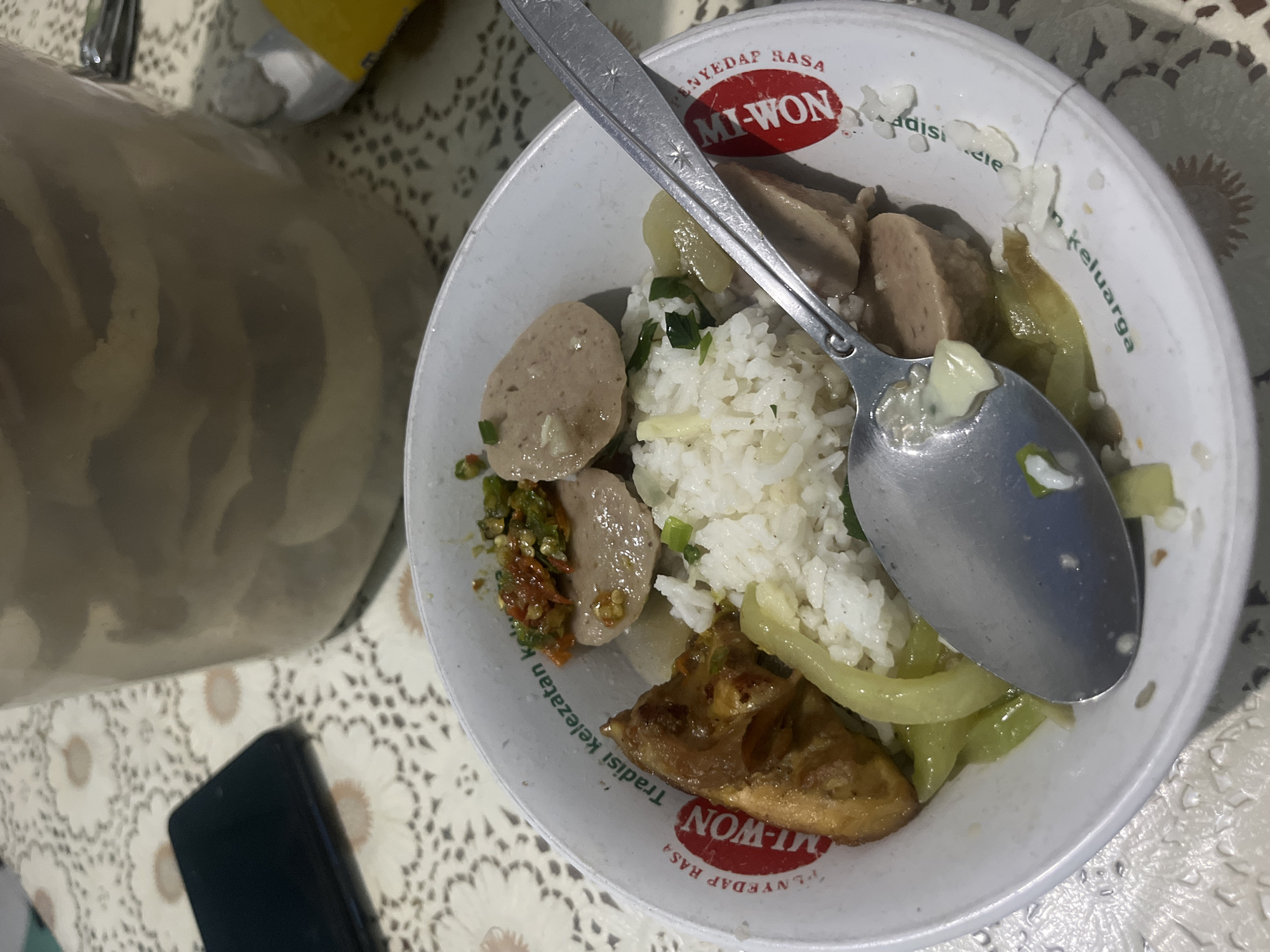 The image shows a bowl of food containing rice, meatballs, vegetables, and a piece of fried tofu. A spoon rests on top of the food. The bowl has the "Mi-Won" brand logo, indicating the use of a seasoning product from the brand. A mobile phone is visible on the table next to the bowl, and the table has a patterned tablecloth. The location, Kranggan, Indonesia, is mentioned in the query.