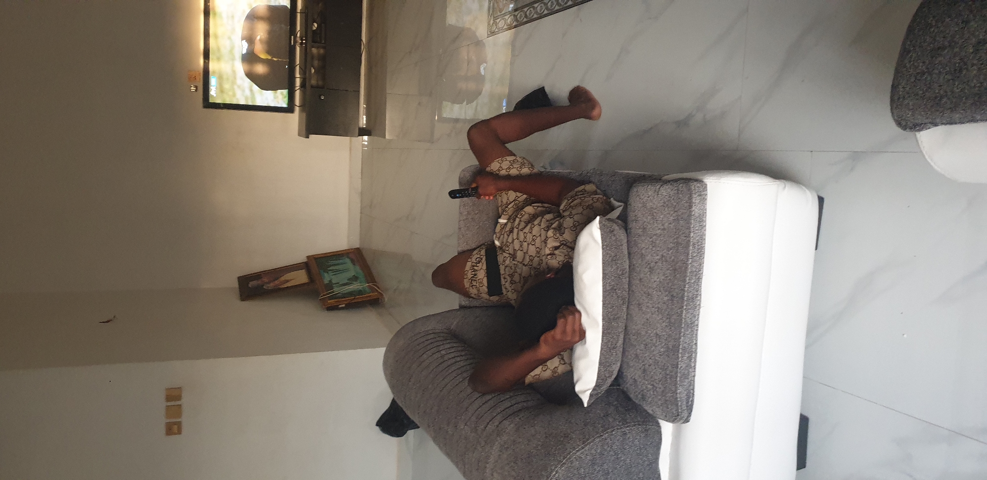 The scene depicts the interior of a residential space located in Narayi, Nigeria. An adult individual with dark skin is positioned on their back on a modern sectional sofa, which features a white base and grey fabric cushions. A grey and white throw pillow supports the person's upper body. The individual is wearing patterned shorts, prominently displaying 'GG' logos and a waistband with 'CHANEL' branding, and is barefoot. Their right hand holds a black television remote control, while their left arm rests on the pillow. The person's legs are bent, with their bare feet positioned over the edge of the sofa.
In the background, a flat-screen television is situated on a dark media cabinet against the far wall. The television screen is illuminated, displaying an image that appears to be a natural outdoor scene featuring an organic form. The flooring throughout the room consists of reflective, light-toned tiles, possibly marble or polished ceramic, exhibiting a subtle veined pattern. A decorative tiled border is visible in the lower right section of the floor. The walls are uniformly light-colored. On the left wall, three identical small, square, light-brown decorative elements are affixed. In the corner of the room, two framed artworks are leaning against the wall; the upper frame contains a vertical portrait, and the lower frame depicts a horizontal landscape, possibly a waterfall. A dark, small object, possibly an insect or debris, is visible on the upper left wall. A dark object, possibly a bag or footwear, rests on the floor near the left side of the sofa.