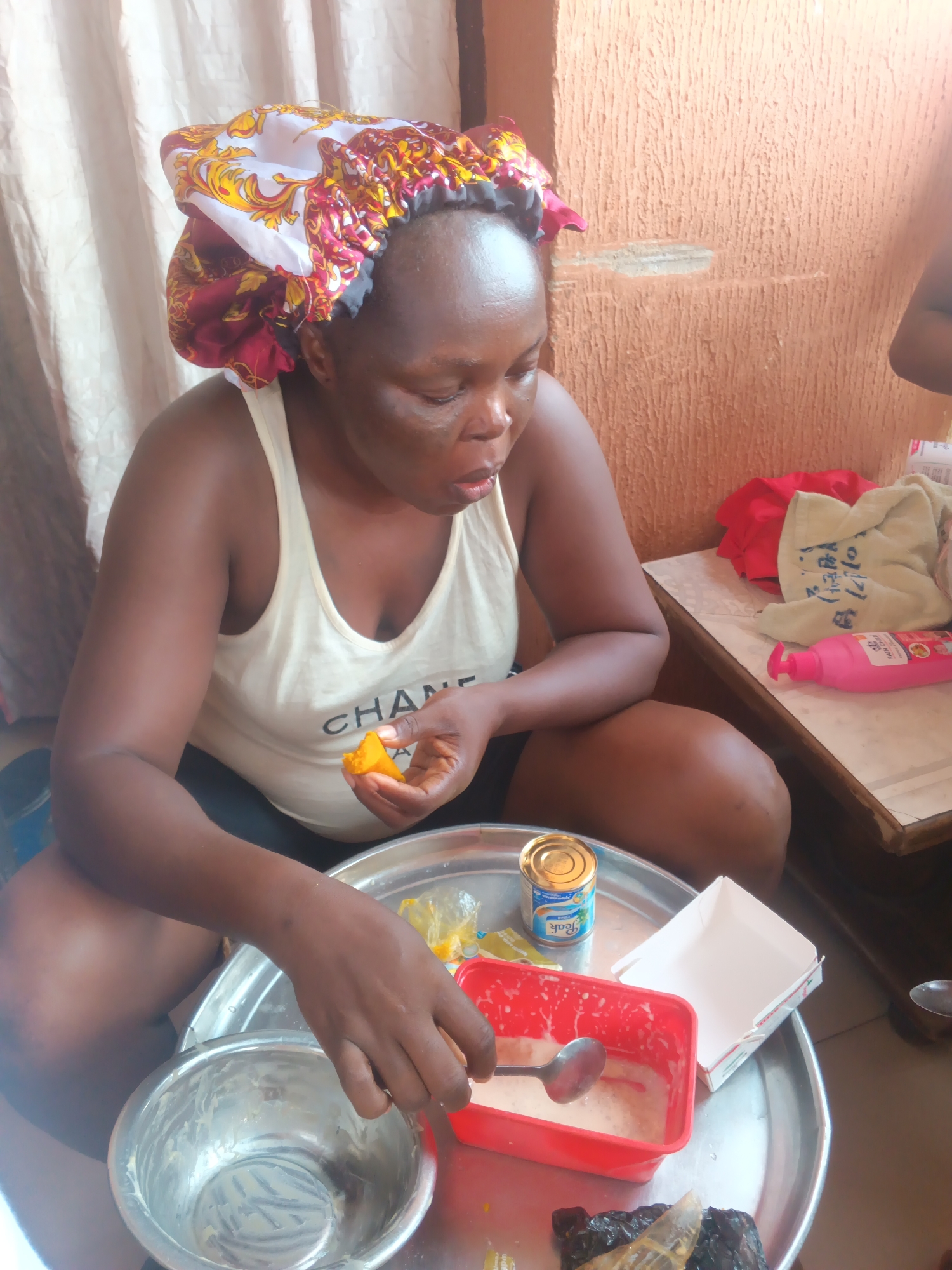 A middle-aged woman wearing a patterned head covering and a white tank top sits cross-legged on the floor. She holds a piece of yellow food in her right hand, bringing it to her mouth. In her left hand, she holds a spoon over a red container filled with a white substance. A silver tray on the floor contains a metal bowl, a can of Peak milk, a white box, and what appears to be dark-colored smoked fish. The background includes beige walls and draped white fabric. The scene is likely indoors, potentially in a domestic setting in Onitsha, Nigeria, given the woman's attire and the presence of Peak milk, a common product in West Africa. The woman appears to be preparing or eating a meal.