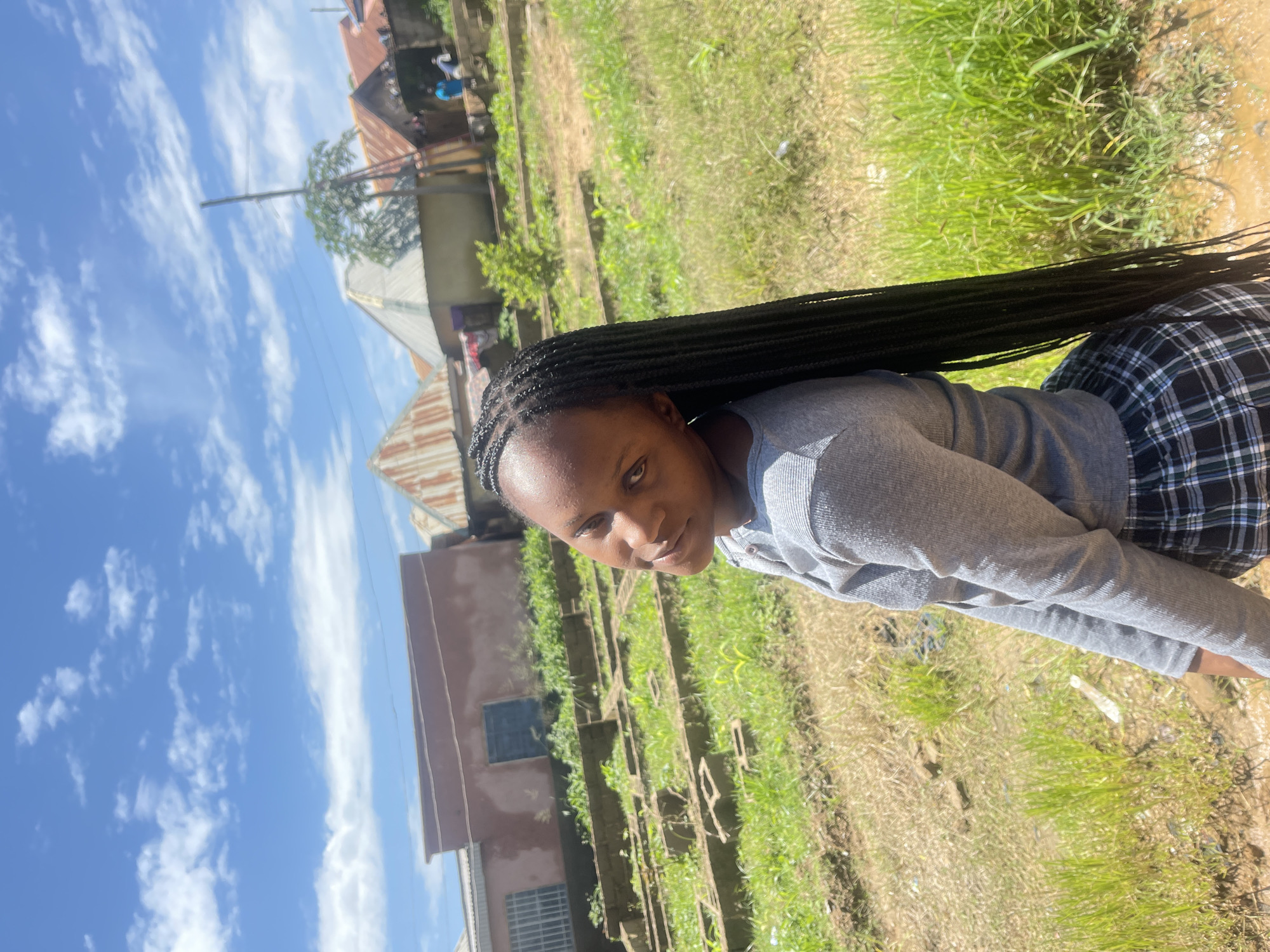 The image is a medium shot of a young Black woman with long braided hair standing outdoors. She is wearing a grey long-sleeved shirt and a black and white plaid skirt. Her head is turned towards the camera, and she is looking slightly to her left with a subtle smile. The background suggests a semi-urban or rural environment in Rafin Par, Nigeria, with buildings that have corrugated iron roofs and a few visible structures that appear to be foundations or garden beds. There is lush green grass and some dry, patchy vegetation in the foreground and middle ground. The sky is blue with scattered white clouds. The lighting indicates it is daytime.