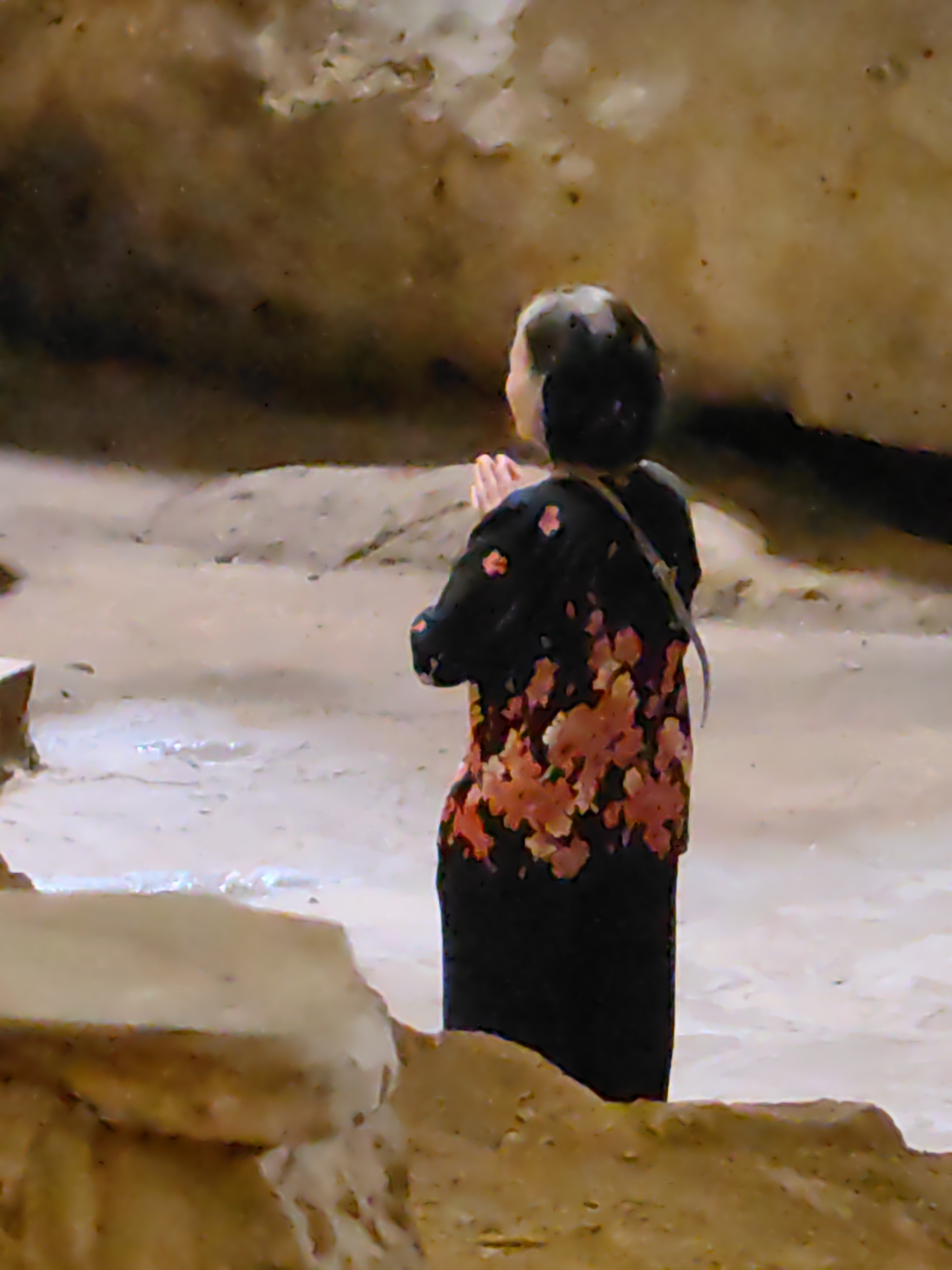 A person stands with their hands clasped in front of them, facing away from the viewer in a sandy, earthy-toned environment. The person has short, dark hair and is wearing a black garment with a floral pattern in shades of orange and red. A thin strap is visible across their back. The surrounding area is rough and uneven, with varying shades of brown and tan suggesting a natural or excavated setting. The location is the city of Lạng Sơn, Vietnam.
