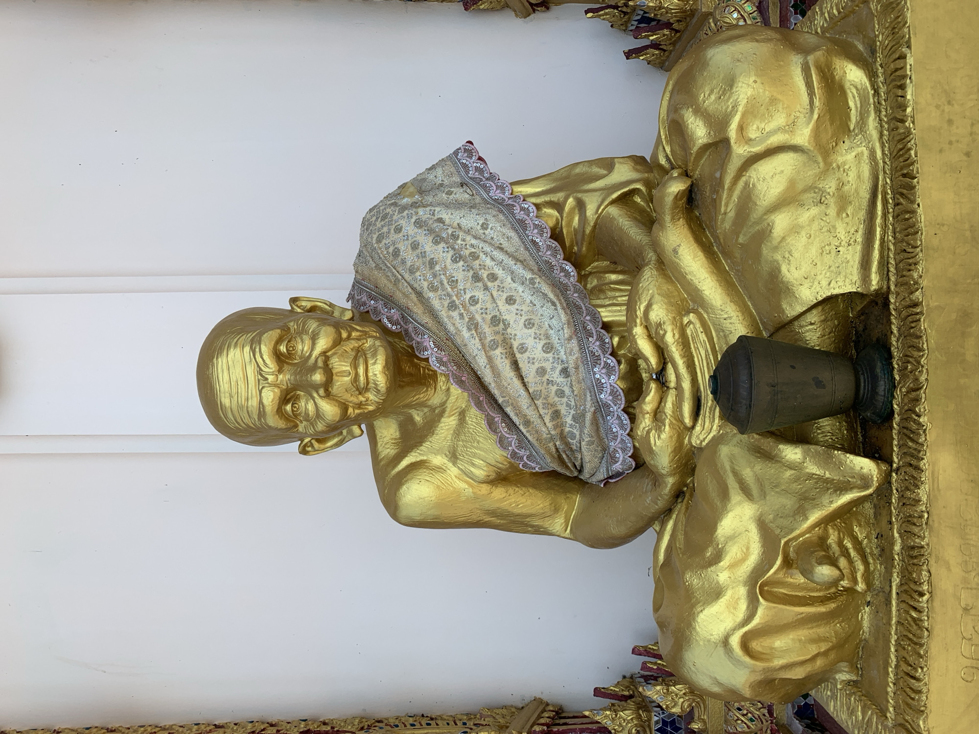 An image depicts a gold-painted statue of an elderly male figure, likely a revered Buddhist monk, seated in a meditative posture on a golden pedestal. The figure exhibits detailed facial wrinkles and hands resting in its lap. A light-colored, patterned fabric with gold and pink embroidered trim is draped over the statue's left shoulder and torso. A dark, metallic-appearing offering vessel rests on the pedestal directly in front of the figure. The statue is positioned against a plain white wall, flanked by colorful, ornate traditional Thai architectural decorations featuring gold, red, blue, and white mosaic-like elements. This scene is located within a temple or religious structure in Baan Plai Laem, Thailand.