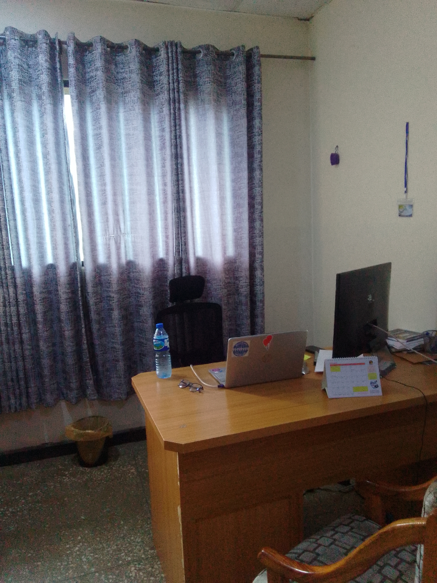 The image depicts an office interior, likely in Jos, Nigeria, characterized by a warm, natural light filtering through patterned grey curtains covering a window. A light brown wooden desk occupies the foreground, holding a laptop, a monitor, a desk calendar, a pair of glasses, and a bottle of water. To the left of the desk, a black office chair is partially visible, and a trash bin with a crumpled liner rests on a speckled floor. The walls are a light, neutral color, with a small purple object hanging on the left and a lanyard with a card holder on the right. The overall impression is of a functional, albeit somewhat dated, office workspace.